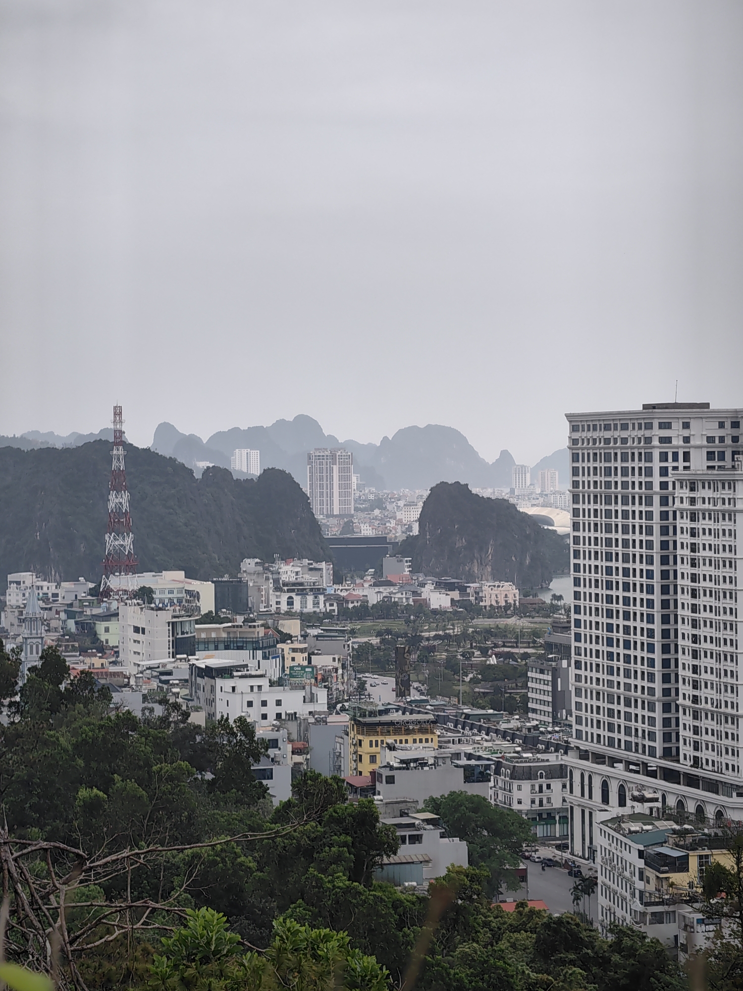This image captures a panoramic, hazy daytime view of an urban landscape set against dramatic karst topography, consistent with Hong Gai Ward, Vietnam.
In the immediate foreground, dark green tree foliage and branches partially obscure the view, suggesting the photograph was taken from an elevated position. The midground showcases a dense urban area featuring numerous multi-story buildings of varied architectural styles. A prominent red and white lattice communications tower stands on a tree-covered hill to the left of the frame, towering over surrounding structures. A large, modern white high-rise building with a grid of windows dominates the right side of the midground. Buildings vary in color and height, including some with green or red roofs, reflecting a mix of residential and commercial spaces.
The background is defined by numerous rounded and jagged limestone karst mountains, characteristic of the region. These mountains appear softened and somewhat indistinct due to a pervasive atmospheric haze or overcast weather conditions, which lend a muted color palette to the entire scene. Between the mountain formations, glimpses of more distant urban development and what appears to be a body of water can be seen.
The weather is overcast and hazy, giving the impression of a subdued daytime. No specific human activity is discernible, as the image focuses on the cityscape and natural environment. Visible text includes "SUN MARINA" on a building in the mid-left, "QBB" on a distant green sign, and "URUGUAY" on a yellow building in the lower-right section. The overall impression is one of a bustling city integrating with its distinctive natural surroundings under a muted sky.