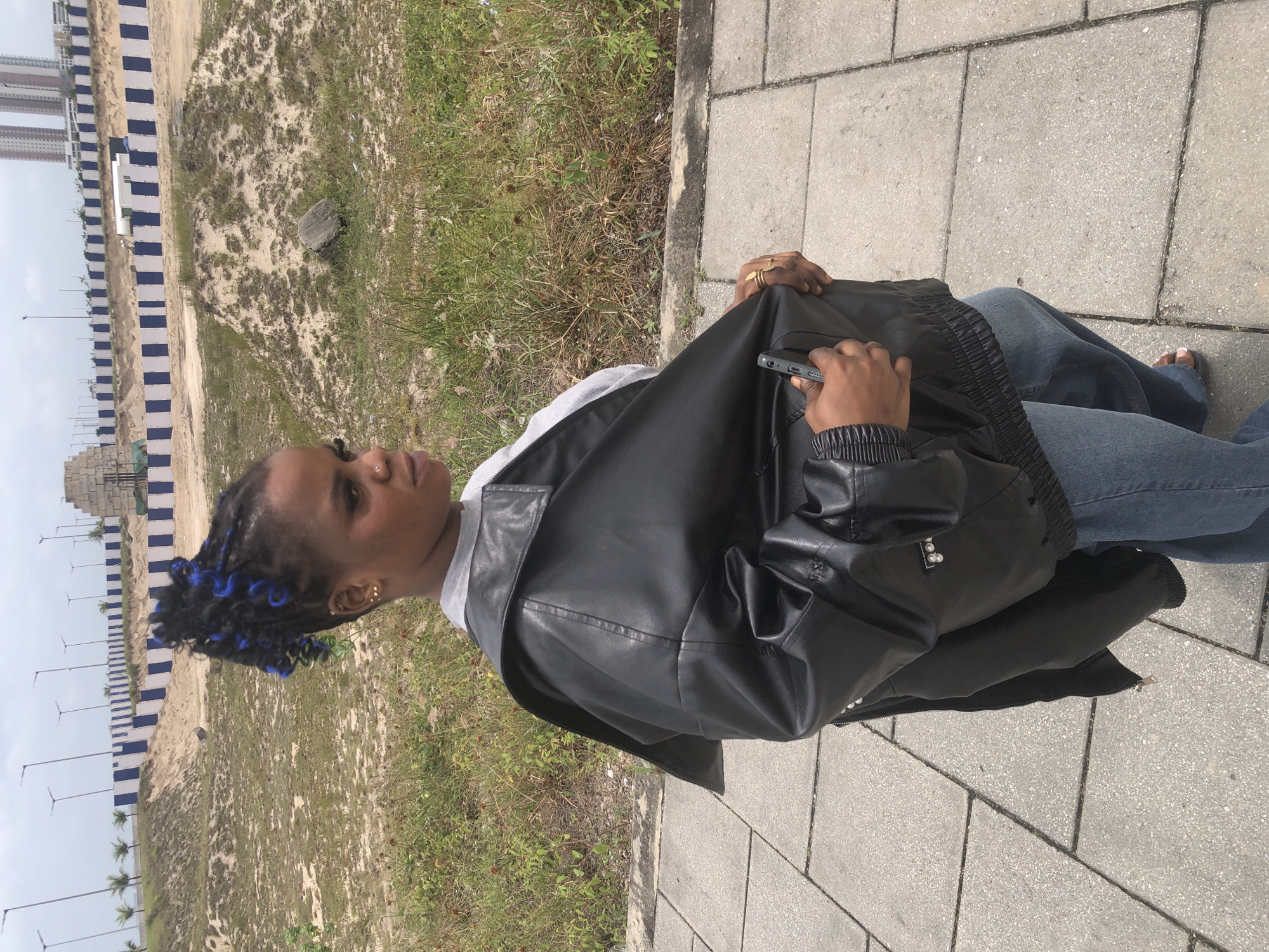 In Ikoyi, Nigeria, a young woman stands on a paved walkway. She is wearing a grey t-shirt, blue jeans, and an oversized black leather jacket. Her hair is styled in braids with blue accents. She is holding a mobile phone in her right hand. The background features a construction site with sand mounds and a fence made of blue and white striped barriers. In the distance, tall buildings can be seen. The sky is overcast.