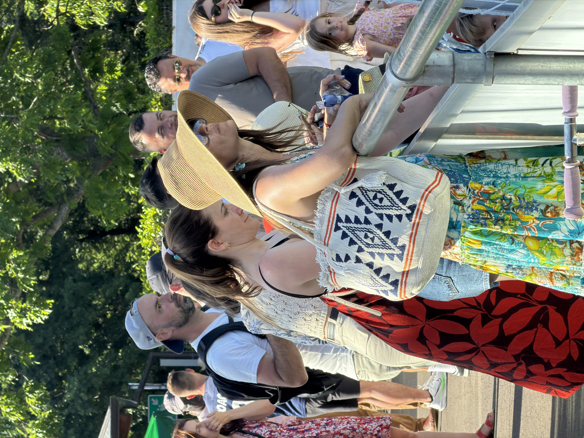 A daytime outdoor scene shows a group of people standing close together in Fentange, Luxembourg. Many of them are looking up and to the left. A woman in the front of the group, facing left, wears a white top and carries a white bag with blue designs. Next to her is a woman in a straw hat, with sunglasses. Above them, others are looking in the same direction, including a man wearing sunglasses. Above that, a child is visible. A metal structure is above the group of people, with metal poles and a white surface. Vegetation, possibly trees, is visible in the background. The people appear to be onlookers at an event.