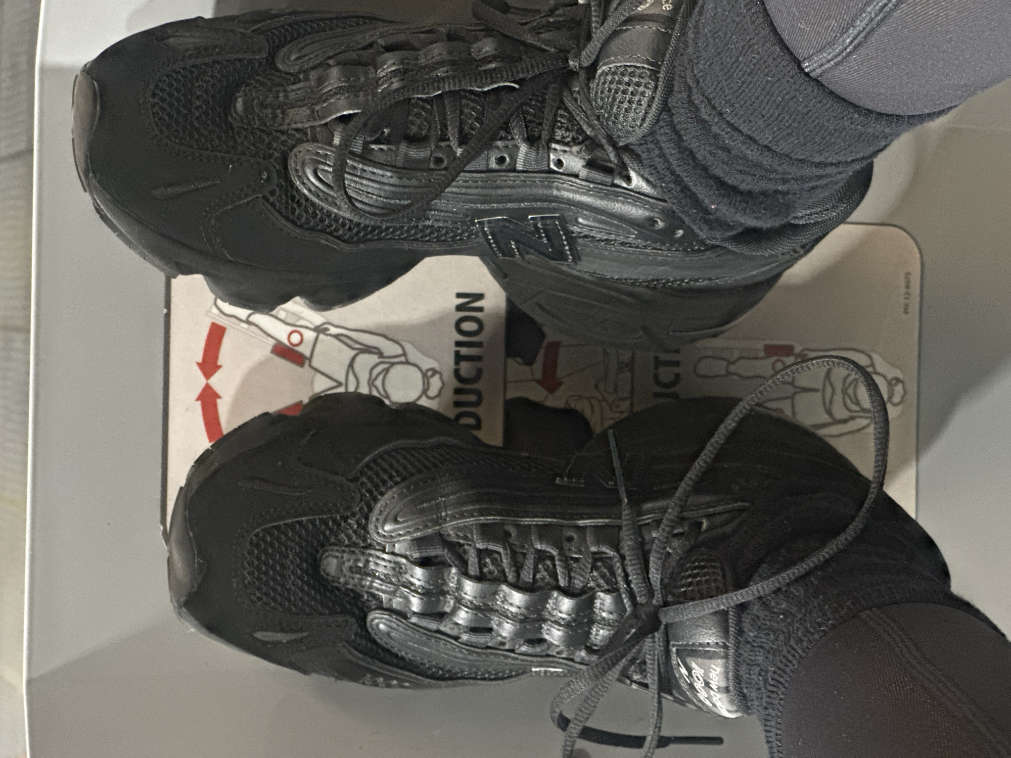 A top-down close-up shot shows a pair of black New Balance sneakers on a white surface with red and black graphics and the word "PRODUCTION" printed on it. The shoes are laced up and appear to be worn with black socks. The surface upon which they rest features a diagram of a person performing a forward bend with arrows indicating direction. The location, Tunbridge Wells, United Kingdom, is not discernible from the visual content of the image.