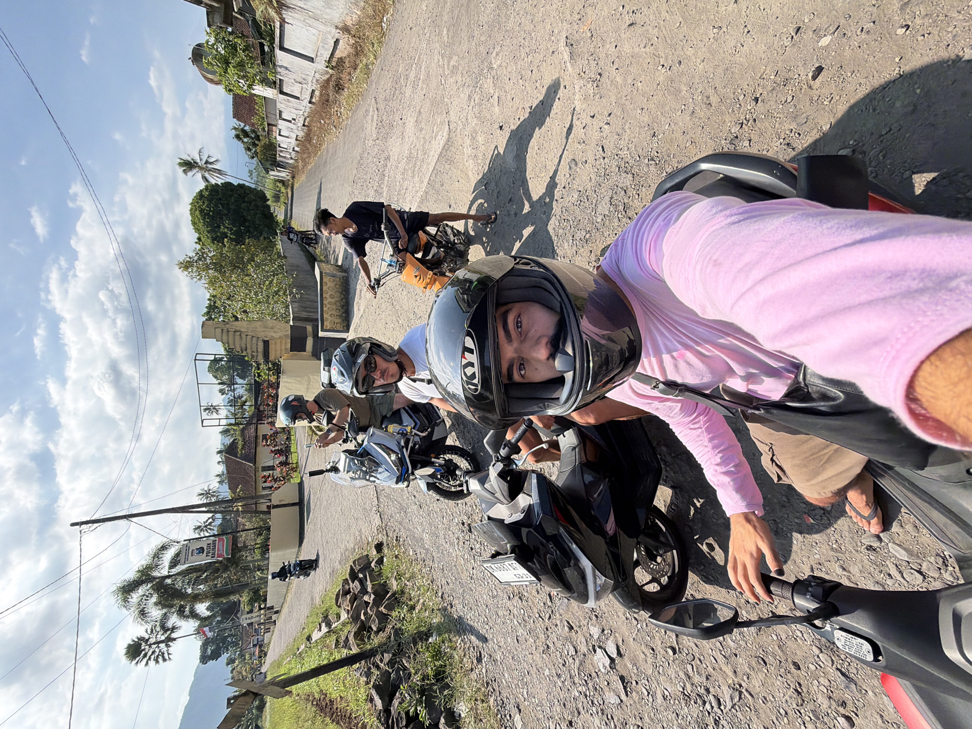 In Kalibaru Kulon, Indonesia, four individuals are positioned on or near motorcycles along a dusty road. The foreground is dominated by a person in a pink long-sleeved shirt and helmet, partially seated on a black motorcycle, holding the handlebars. To their left, another person in a green shirt and helmet is on a white scooter, holding a phone. Behind them, two other individuals are also on motorcycles. The road is unpaved and surrounded by vegetation and buildings. A banner with Indonesian text and a police emblem is visible on a pole to the left, further identifying the location. The sky is partly cloudy, and the overall scene suggests a warm, sunny day.