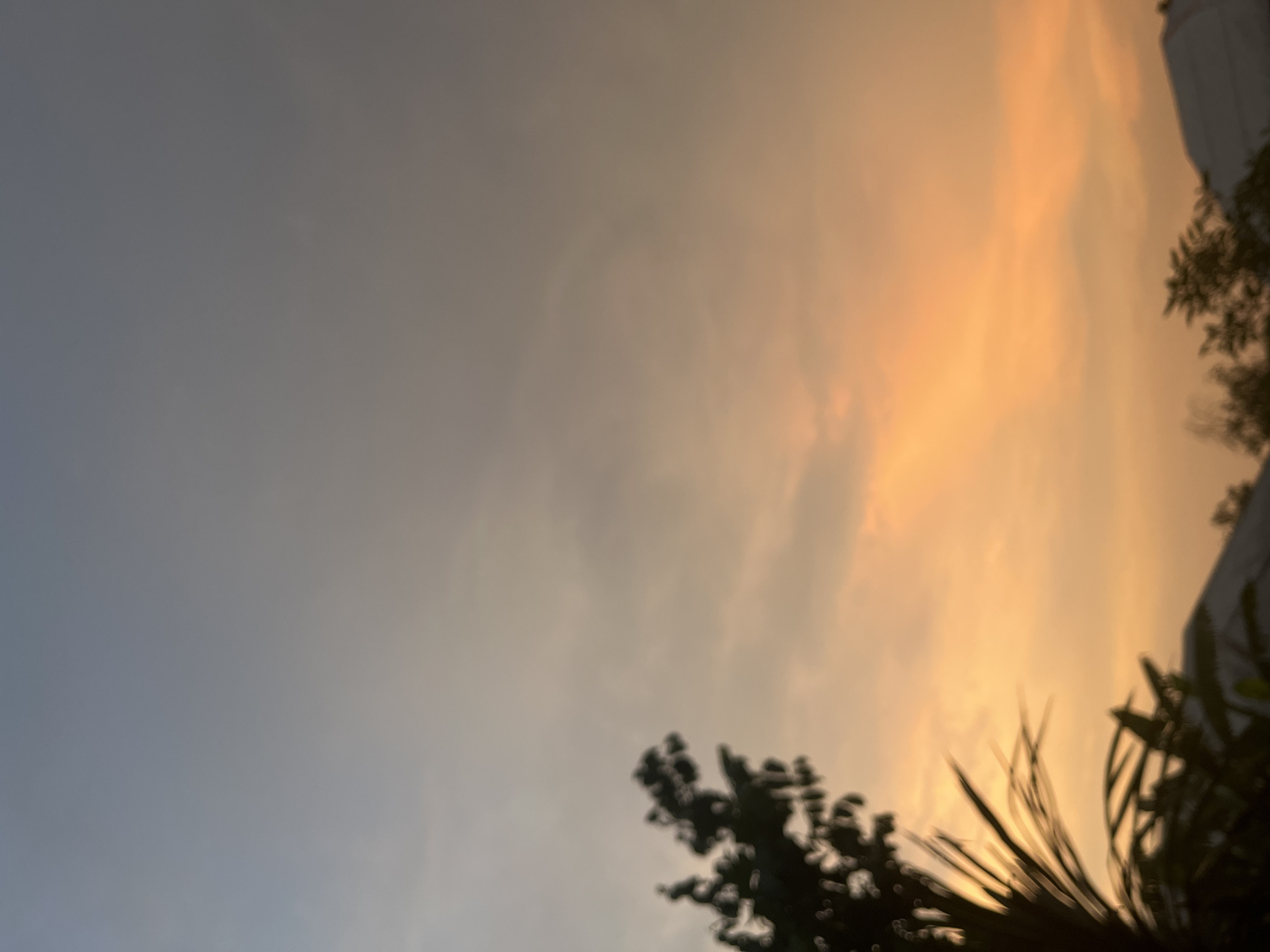 The image shows a sky at dusk in what may be Ikot Ekpene, Nigeria. The sky transitions from a greyish-blue on the left to orange on the right, suggesting a sunset or sunrise. Silhouetted trees and possibly a building are visible against the colorful sky. The focus appears to be soft, resulting in a slightly blurry image.