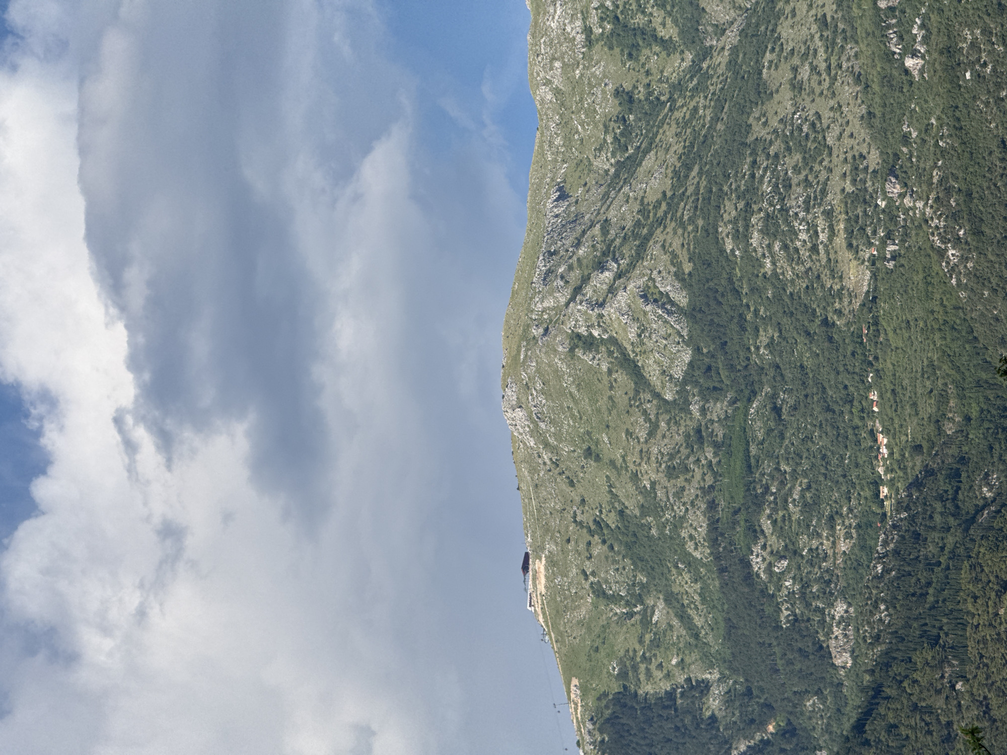 The image shows a view of a steep, green mountainside with a blue sky and clouds above. The mountain is covered in vegetation, with patches of lighter rock visible. There are some small buildings nestled into the hillside. In the distance, a cable car is visible, ascending the mountain. The scene suggests a sunny day with some cloud cover. The overall impression is one of natural beauty and a recreational activity.