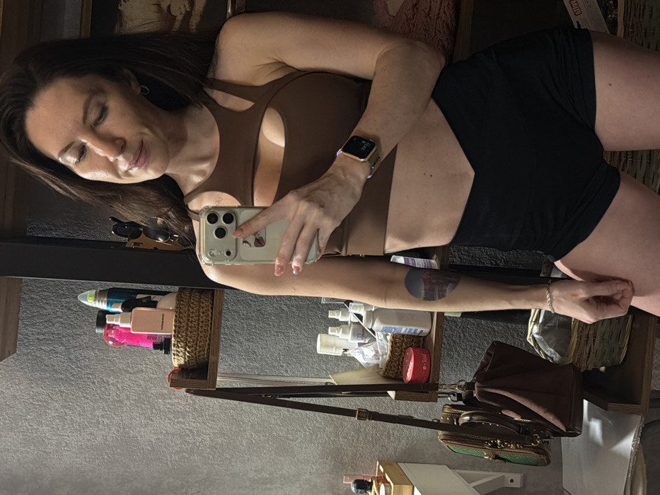 A woman in a brown top and black shorts poses for a selfie. She holds a phone in her right hand, wearing a smartwatch on her left wrist. Her hair is brown and shoulder-length. Shelves with various toiletries and a brown handbag are visible in the background. The image appears to be taken in front of a mirror.