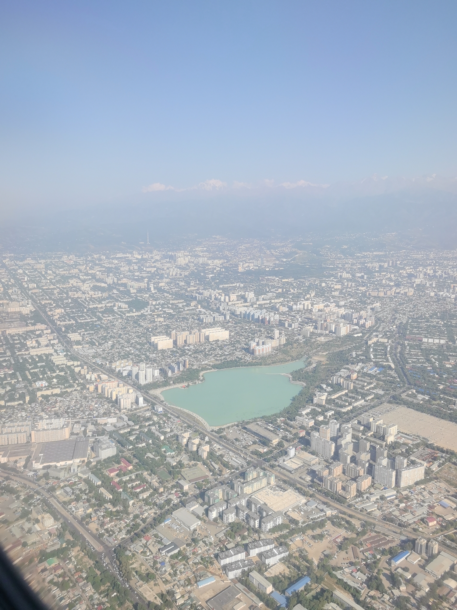 An aerial, wide shot captures a sprawling urban landscape under a clear, pale blue sky. The city stretches out towards a hazy horizon where distant, snow-capped mountains are faintly visible. The foreground is dominated by a large, irregular-shaped body of turquoise water, appearing as a lake or reservoir, with its edges lined by verdant trees and a modern cityscape. The city itself is a dense grid of buildings, ranging from low-rise residential blocks to taller apartment complexes and office buildings, interspersed with roads, green spaces, and smaller industrial or commercial structures. The overall impression is one of a densely populated, modern city nestled against a backdrop of natural mountain scenery.