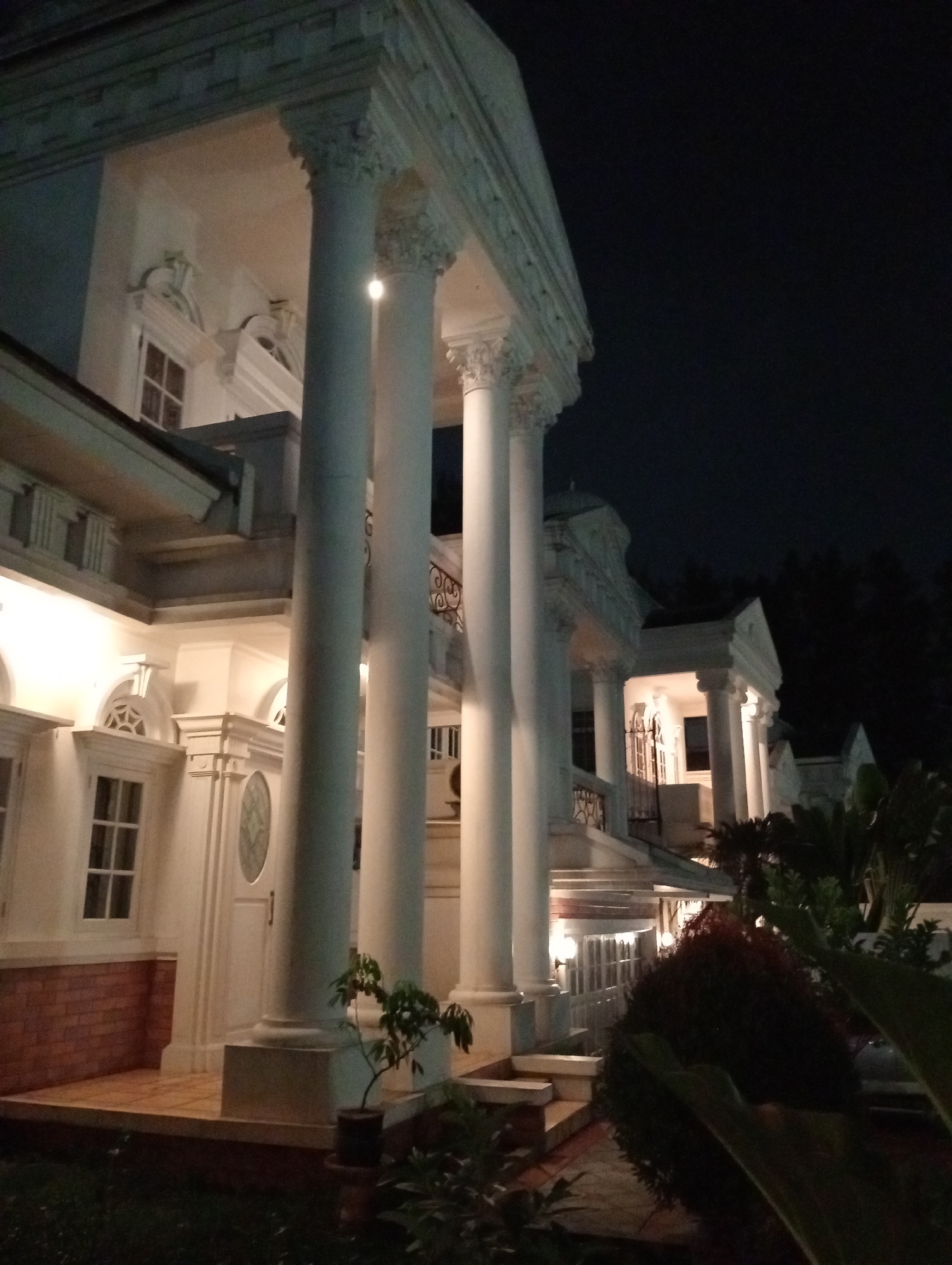 A wide, low-angle shot captures the illuminated facade of a large, white, classical-style building at night. The building features tall, fluted columns supporting an entablature, with detailed architectural elements like cornices and decorative reliefs. Multiple windows, some with grids and arched tops, are visible, along with a prominent doorway with a circular, frosted glass insert. A red brick base is present on the lower portion of the structure. The scene is illuminated by artificial lights, casting a warm glow on the white surfaces and creating shadows. Lush, dark green foliage and trees frame the right side and background of the image, contrasting with the bright building. A small potted plant sits on a wooden porch in the foreground. The sky above is dark and clear. The image appears to be taken from a low vantage point on a lawn.