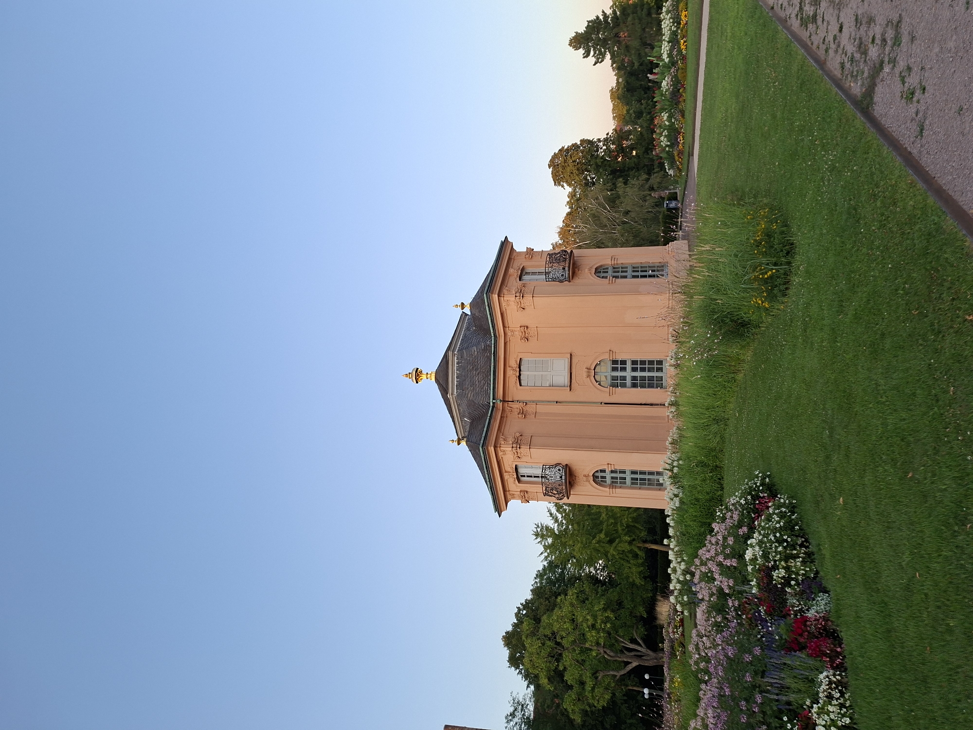 A light pink octagonal building with a dark roof topped with gold finials stands in a park. The building has multiple windows, some arched and some rectangular, and two balconies with decorative railings. The building is surrounded by lush green grass and flower beds filled with a variety of colorful flowers, including pink, white, and red blooms. Trees with dense green foliage are visible behind the building and on the sides. The sky above is clear and a pale blue. The building is the Favorite Palace in Rastatt, Germany.