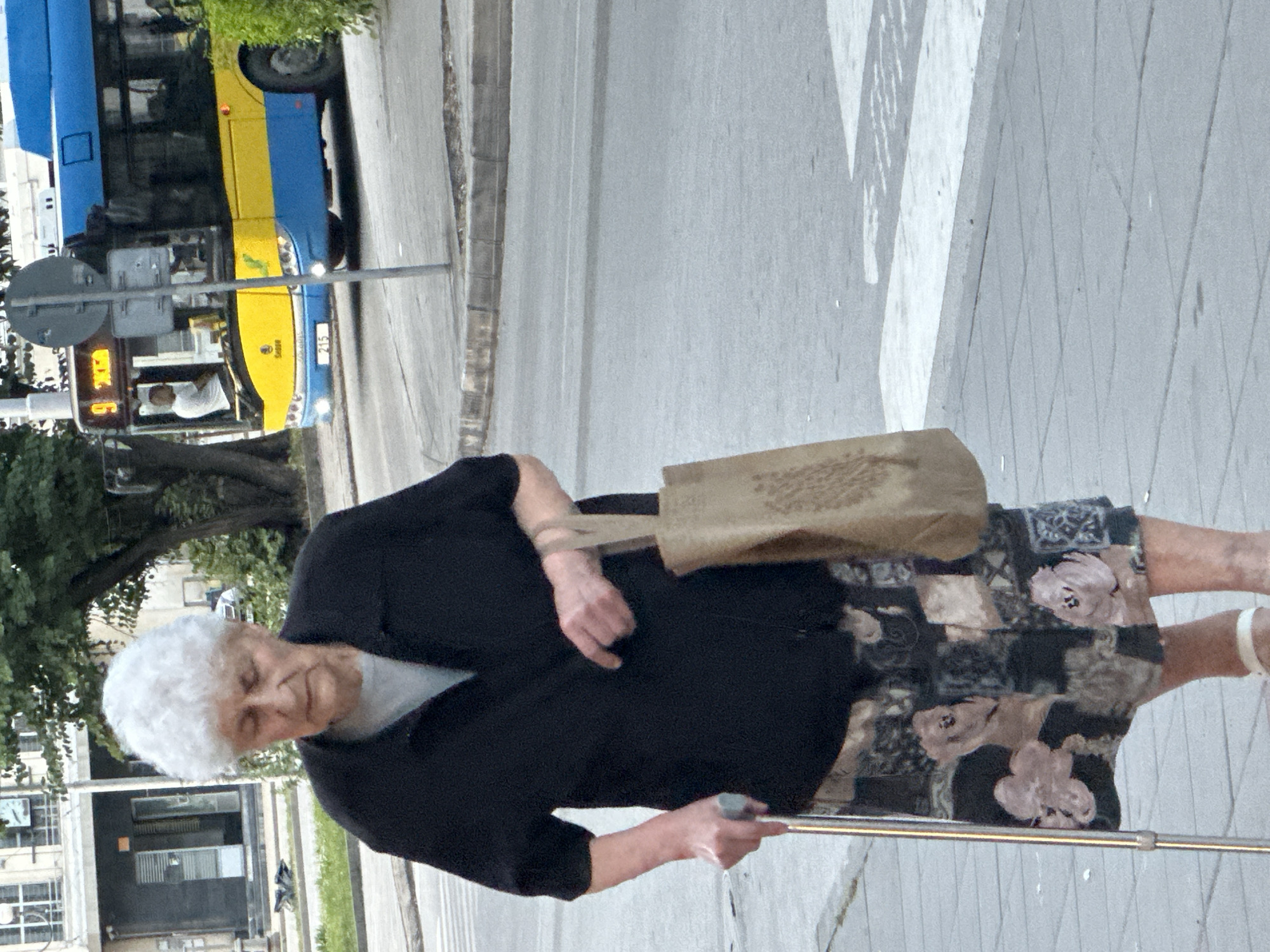 An elderly woman with white hair is walking on a sidewalk in Pleven, Bulgaria, holding a cane in her left hand and a shopping bag in her right. She is wearing a black short-sleeved shirt over a light-colored t-shirt and a patterned skirt. A blue and yellow bus is visible in the background, stopped at a traffic light on a street. The woman is walking from left to right across a crosswalk.