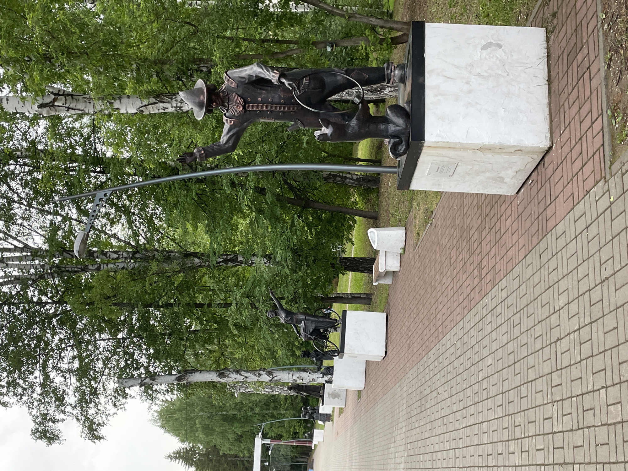 The image shows a series of bronze statues on concrete plinths along a pedestrian walkway. The walkway is paved with two colors of rectangular blocks, red-brown and light gray. The statues depict human figures, and at least one appears to include an animal. Trees with white bark are visible in the background, and green foliage covers the trees. A bench is located behind one of the statues. The image depicts the monuments on the embankment of the Kama River in the city of Perm, Russia.