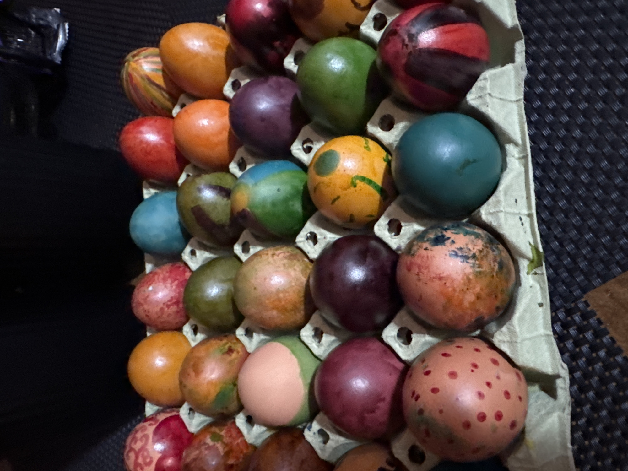 An eye-level close-up shot depicts a carton of twenty-eight decorated Easter eggs. The eggs vary in color and design, including solid colors, patterns, and textures. The carton is made of light-colored, possibly recycled cardboard with individual egg compartments. The background is a dark, textured surface. The image was likely taken indoors. There is no information suggesting that the image was taken in Sofia, Bulgaria.