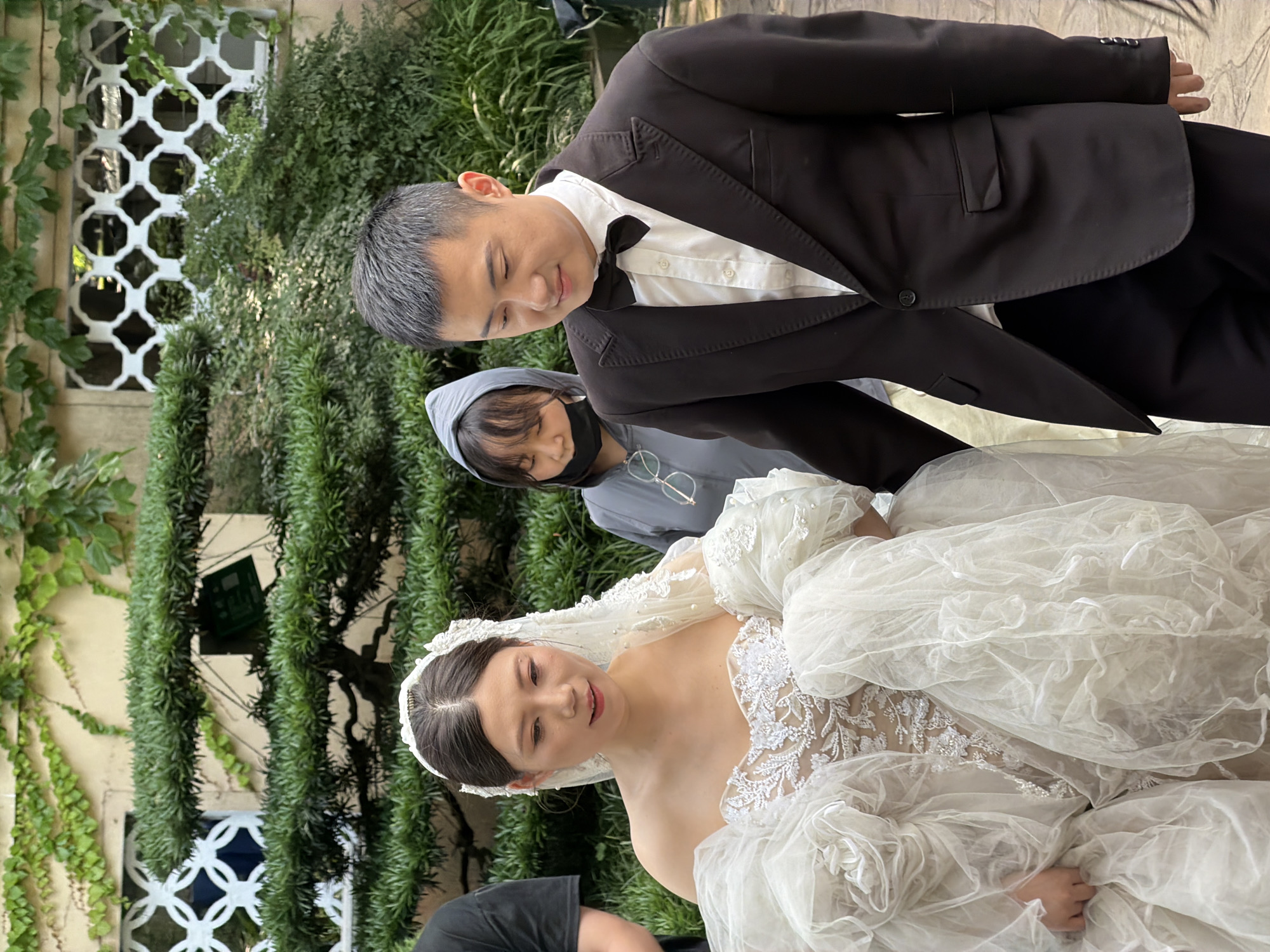 In the Yuzhong District of China, a bride in a voluminous white wedding gown stands next to a groom in a black suit with a white shirt and black bow tie. The bride wears a veil adorned with lace and pearls. Behind them, a woman in a grey hooded jacket and a black face mask is visible. The background features lush green foliage and architectural elements, including a wall with decorative white latticework. The scene appears to be outdoors, possibly in a garden setting, with natural light illuminating the subjects.