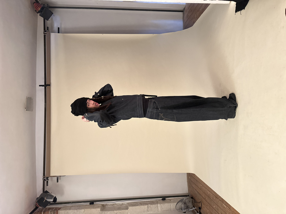 The image captures a single individual posing in a photography studio. The subject, appearing male, stands in a partial profile facing right, with their head turned to look over their right shoulder towards the viewer. They have long, dark dreadlocks partially visible under a black, textured turban-style beanie. The individual is dressed in a dark, possibly indigo, denim jacket with buttoned cuffs, worn over a dark undershirt, and very wide-leg, dark denim jeans featuring distinct stitching on the back pockets. Dark footwear is visible. Both hands are raised near their ears, fingers slightly splayed, in an apparent gesture of adjusting their headwear or hair. Their mouth is slightly open.
The background is dominated by a seamless light beige photographic backdrop that curves from the vertical wall to the horizontal floor. To the left of the backdrop, a section of white-painted exposed brick wall is visible, along with a chrome-colored, cage-covered floor fan on a light-colored wooden floor. To the right of the backdrop, a segment of a plain white wall and more of the light-colored wooden floor are seen, with a crumpled dark fabric item on the floor. Overhead, two black studio lights equipped with barn doors are mounted on a horizontal rail system, positioned to illuminate the subject from both sides. This scene takes place within a photography studio, presumed to be in Paris, France.
