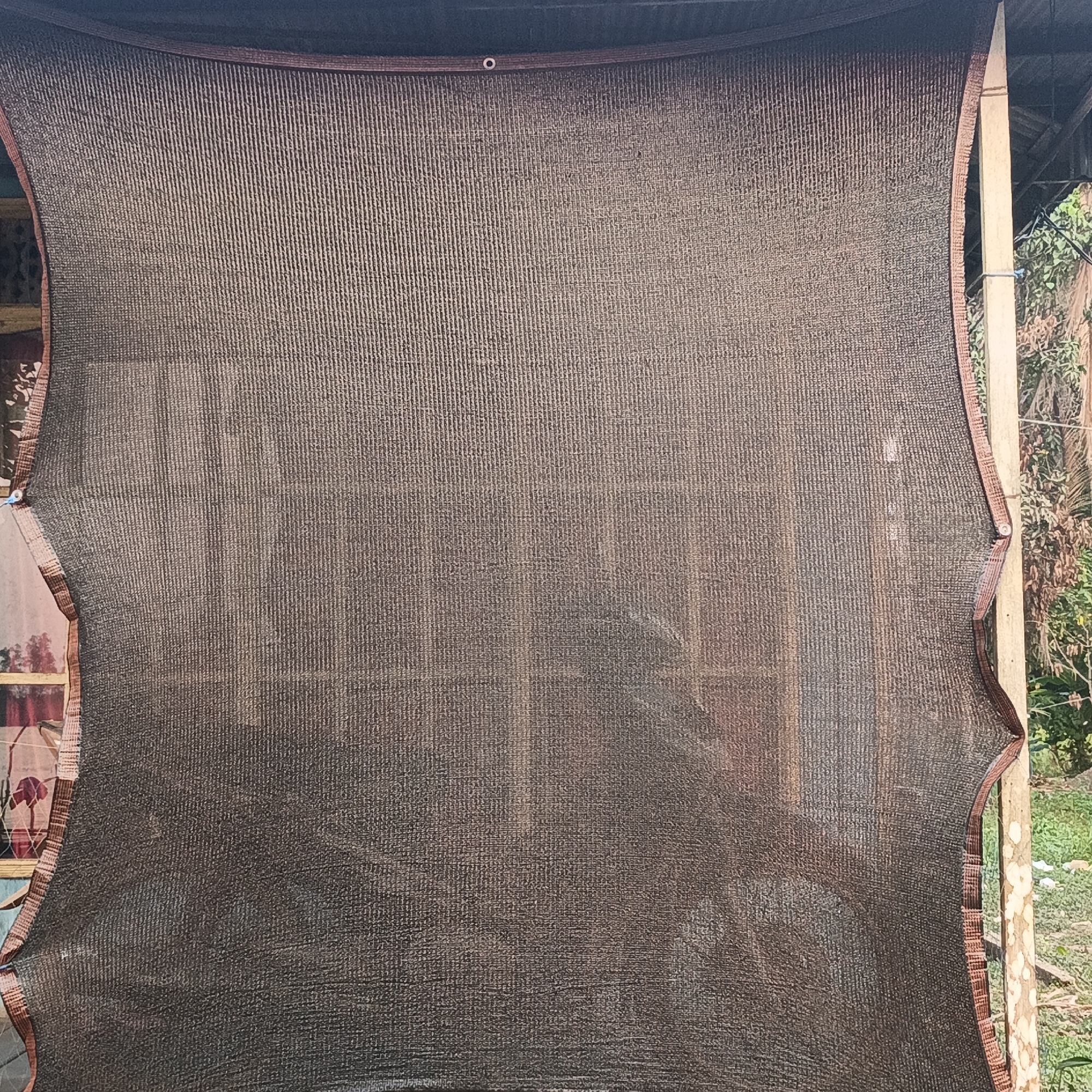An outdoor shot captures a large piece of brown shade cloth tautly stretched between two vertical wooden posts, forming a barrier or covering. The shade cloth is a tightly woven mesh with a visible vertical ribbed texture. It is bordered along its edges by a darker brown trim, which appears to be reinforced with stitching. The top edge of the cloth has grommets, suggesting it's meant to be secured with ties or fasteners.
Behind the shade cloth, a wooden structure is partially visible, characterized by horizontal beams and vertical supports. Through the mesh, faint outlines of what appear to be windows with curtains can be discerned on the left side. The background on the right shows a natural environment with green foliage and trees.
The scene is outdoors, with ambient light suggesting it is daytime. The overall impression is that of a functional outdoor setup, possibly for shade or privacy, in a semi-rural or residential setting. The location is identified as Sungai Rasau, Indonesia. There are no people visible in the frame.