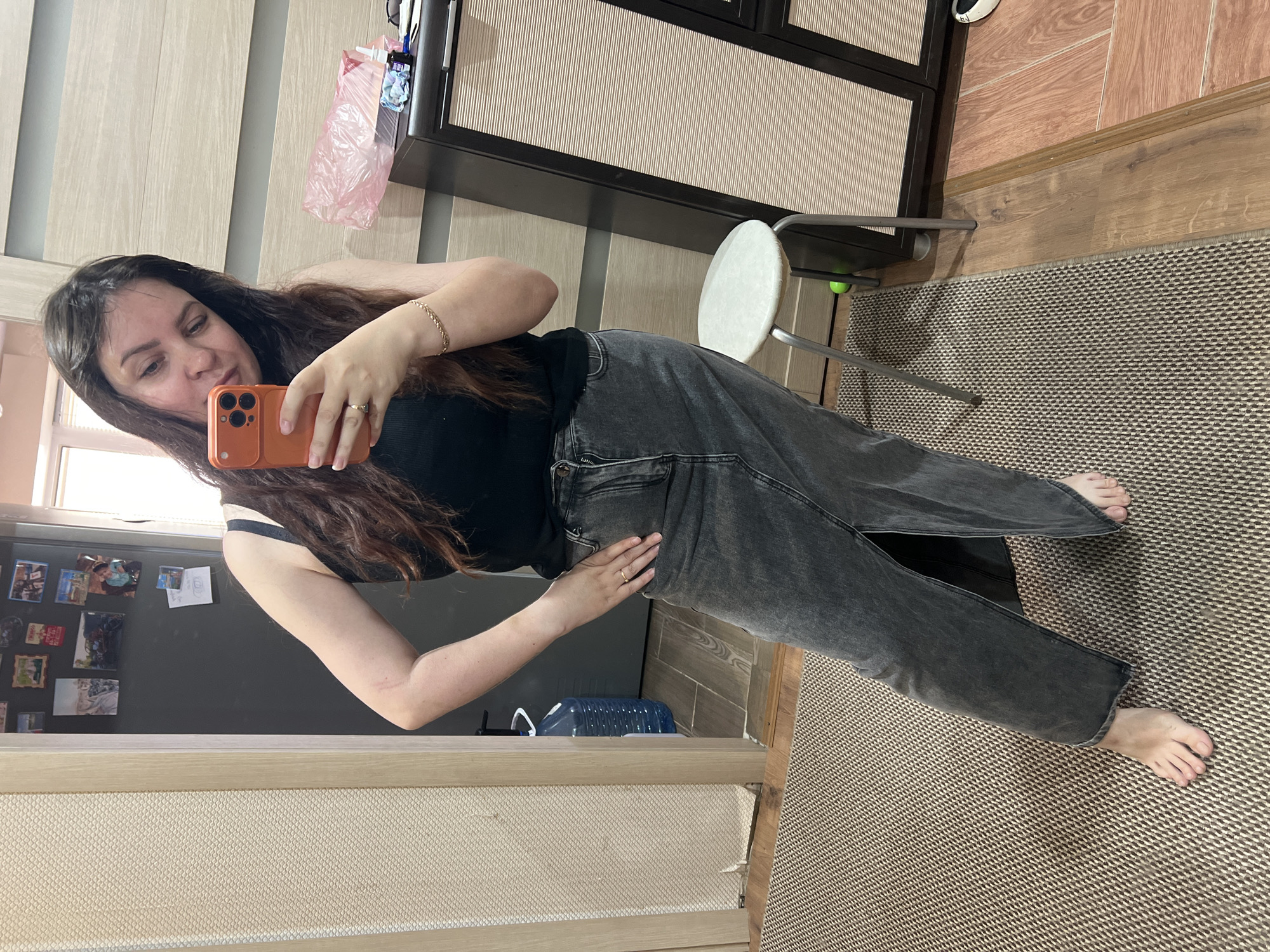 A young woman with long, dark brown hair takes a mirror selfie indoors. She is dressed in a black tank top and dark grey, loose-fitting jeans, and is barefoot. She holds an orange smartphone in her left hand, looking at its screen, while her right hand rests on her hip. She wears a gold-colored bracelet on her right wrist and a ring on her left ring finger.
The setting is a residential interior, suggested to be in Noviy, Russia. The background features various elements: on the left, a light-colored wooden door or cabinet; on the right, a darker brown cabinet with textured light-colored panels. A white round-seated stool with a metal frame is positioned near the dark cabinet. The floor transitions from light wooden planks to a rug with a beige and brown geometric pattern.
Behind the woman, to her left, a dark surface, likely a refrigerator door, is decorated with several photos and magnets. A blue plastic bottle rests on the floor. A pink plastic bag hangs in the upper right portion of the frame. The scene is illuminated by natural daylight. The woman's expression is neutral and focused on the phone. No visible text is present in the image.