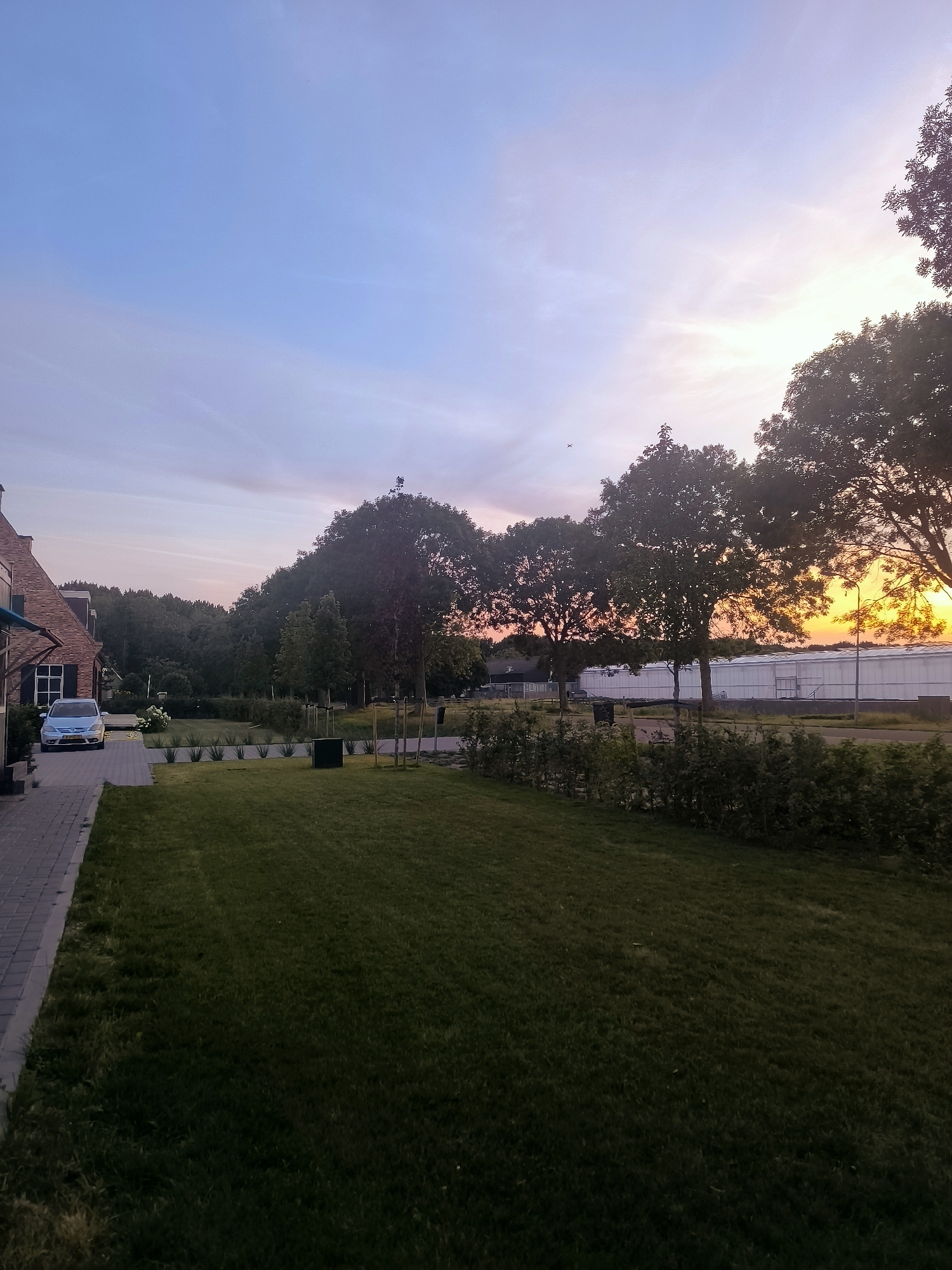 The image shows a sunset scene in Almere, Netherlands. In the foreground, there is a neatly mowed lawn and a brick pathway leading to a house. A silver car is parked on the pathway. In the middle ground, there are several trees and a low hedge. In the background, there are large greenhouses and more trees. The sky is a gradient of blue, pink, and orange, indicating the sun is setting. The overall scene depicts a suburban or semi-rural environment.