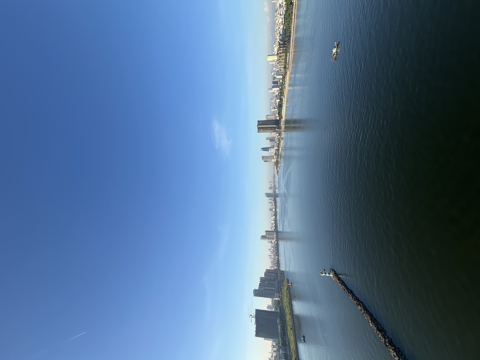 An aerial, wide-angle shot captures a serene panoramic view of the Han River in Da Nang, Vietnam, under a clear, bright blue sky with a faint contrail from an airplane. The river, a dominant feature, flows from the upper left to the lower right of the frame, reflecting the clear sky. On the left bank, a cluster of modern high-rise buildings, some under construction with visible cranes, stands prominently. Along the riverbank, a breakwater, constructed of dark rocks, extends into the water, topped by a small lighthouse with white and green markings. Further into the river, several small boats are visible. On the opposite bank, a long stretch of urban development lines the river, featuring a diverse array of buildings, from residential structures to more contemporary skyscrapers, stretching into the distance. The scene is tranquil, with no visible people, suggesting an early morning or late afternoon setting.
