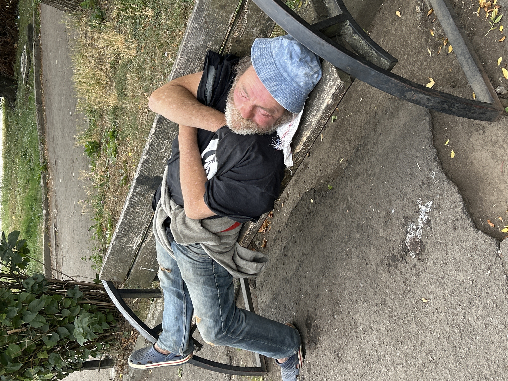 An adult male is sleeping on a park bench. He is wearing a black t-shirt, blue jeans, and a blue hat. The bench is made of wood and metal, and it is located outdoors in a park. There is a bush and grass in the background, and the ground is paved. The person appears to be homeless. The scene is in Uzhhorod, Ukraine.