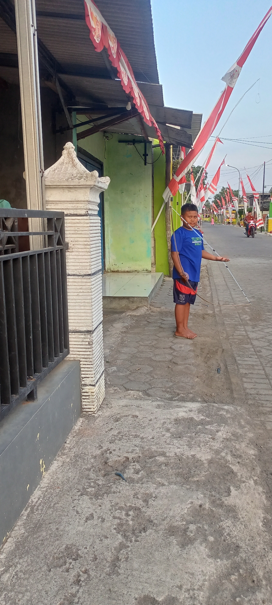 The image shows a boy in Gempolklutuk, Indonesia, standing on a paved street. He is wearing a blue t-shirt and patterned shorts and is holding a long pole that extends to the right, with a string attached. The street is lined with red and white flags, typical of Indonesian national celebrations. A green building with a corrugated metal roof is visible behind the boy, and a black metal fence runs along the left side of the image. In the background, other flags are strung across the street, and a person is seen riding a motorcycle. The ground is a mix of concrete and loose dirt. The lighting suggests it is late afternoon or early evening.