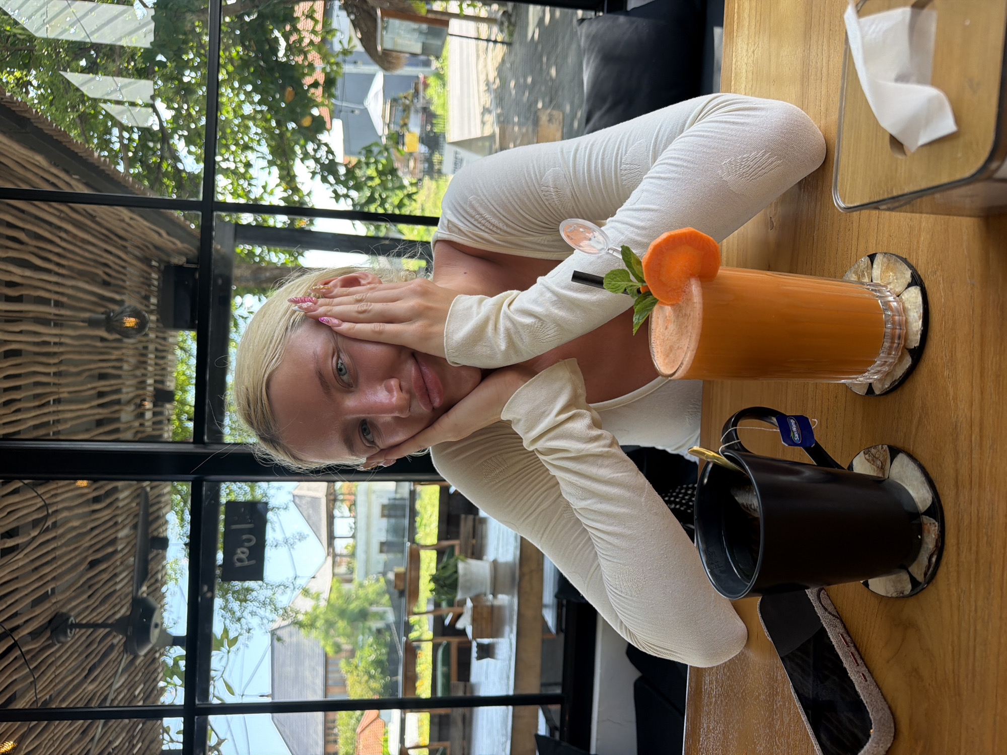 A young Caucasian woman with blonde hair is seated at a wooden table in what appears to be an open-air cafe in North Kuta, Indonesia. The woman is leaning her head on her hands, with her elbows resting on the table. She is wearing a long-sleeved, cream-colored top with a textured pattern. In front of her, there are two drinks: a black mug of tea with a tea bag and a spoon, and a tall glass of orange juice garnished with a slice of carrot and a mint sprig. A smartphone with a grey case is placed to the left of the mug. In the background, through large windows, a street scene with buildings, trees, and parked vehicles is visible. The cafe has a rustic ceiling made of natural wood branches and a ceiling fan. The overall atmosphere suggests a casual dining or relaxation setting.