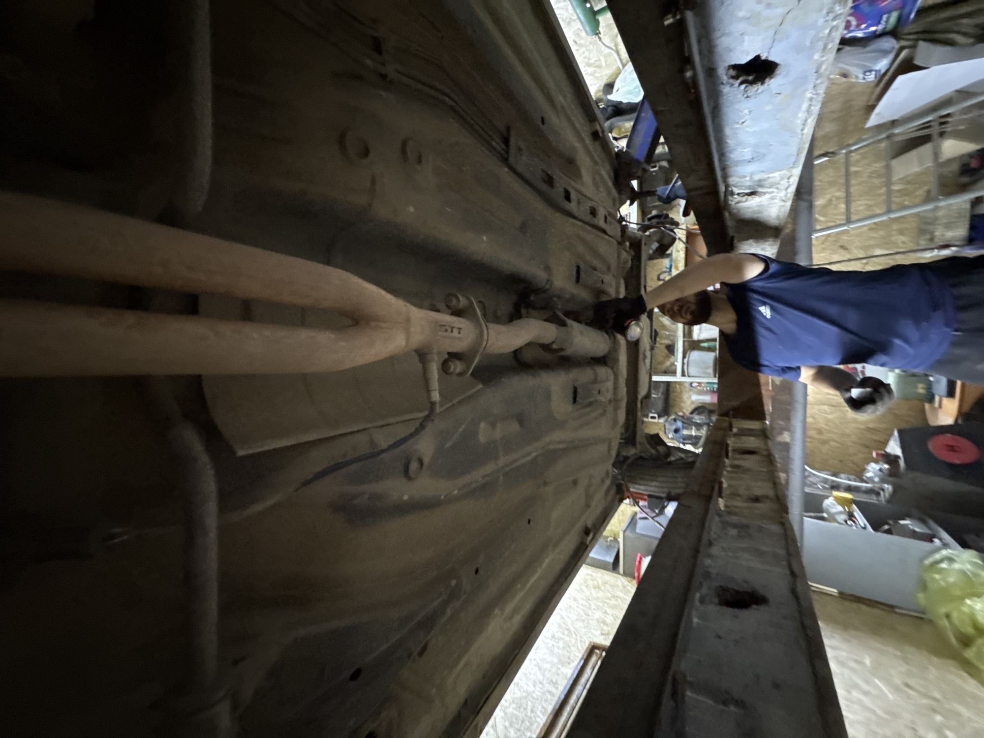 This image presents a low-angle perspective looking upwards at the underside of a car, situated within a workshop environment in the city of Perm, Russia. A male individual, characterized by a beard and wearing a dark blue t-shirt with an Adidas logo and dark gloves, stands beneath the vehicle. He holds a metallic flashlight in his raised right hand, directing its beam toward the car's undercarriage.
The car's underside exhibits visible dirt and areas of surface rust. A dual-pipe exhaust system, merging into a single pipe, is prominent, with a visible flange connection marked "STT". The vehicle's chassis, floor pan, and various structural components are discernable. The car is supported by a heavy-duty, rust-stained metal lift or support structure, which features multiple rectangular cutouts along its length. A blue hydraulic jack or similar support device is positioned on the right side of the scene.
The surrounding workshop features walls clad in light-colored wooden paneling, possibly oriented strand board (OSB). The background contains various objects typical of a garage, including a silver metal ladder, shelves, tools, containers (one red), and plastic bags. The lighting appears to be artificial, illuminating the workspace.