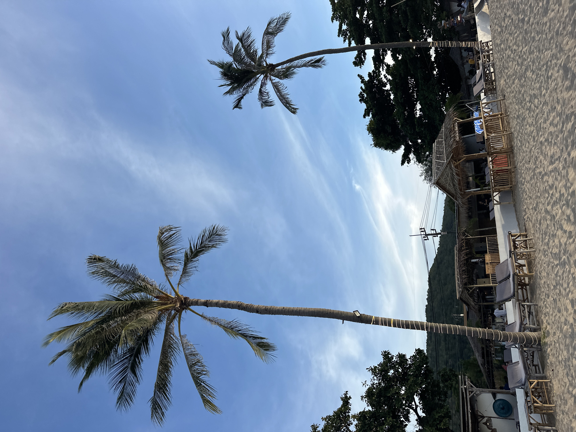 The image depicts a sandy beach scene in the Ko Samui District of Thailand. Two tall palm trees are prominently featured against a backdrop of a light blue sky with wispy white clouds. The foreground is a sandy beach with several lounge chairs and small bamboo tables set up. Behind the chairs, there is a beachfront establishment with thatched-roof structures, possibly a bar or restaurant, and some customers are visible. Lush green trees and foliage line the background, and in the distance, hills can be seen, with a few utility poles and wires crossing the sky. The overall scene suggests a relaxed, tropical atmosphere.