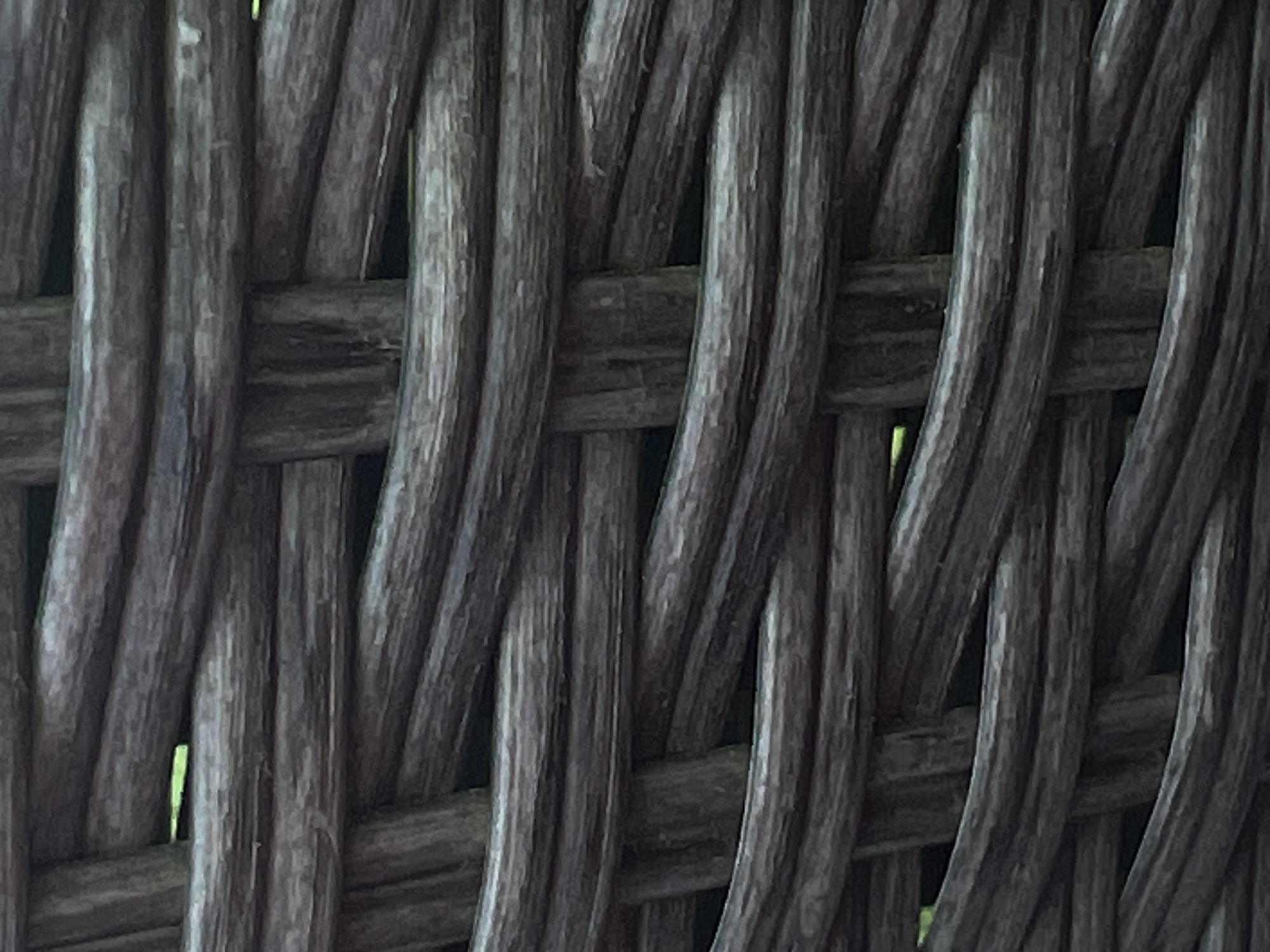 The image displays a close-up view of woven, dark brown material, likely rattan or wicker. The texture is coarse and the individual strands are thick and rounded. The weaving pattern is a simple over-under interlacing. In some of the gaps between the woven strands, a small sliver of green is visible, suggesting an outdoor environment or a backdrop of foliage. The lighting is subdued, creating shadows and highlighting the texture of the material. There are no people or any discernible objects beyond the woven material itself. The image does not contain any specific landmarks or architectural features that would allow for the identification of a precise location within Genemuiden, Netherlands.