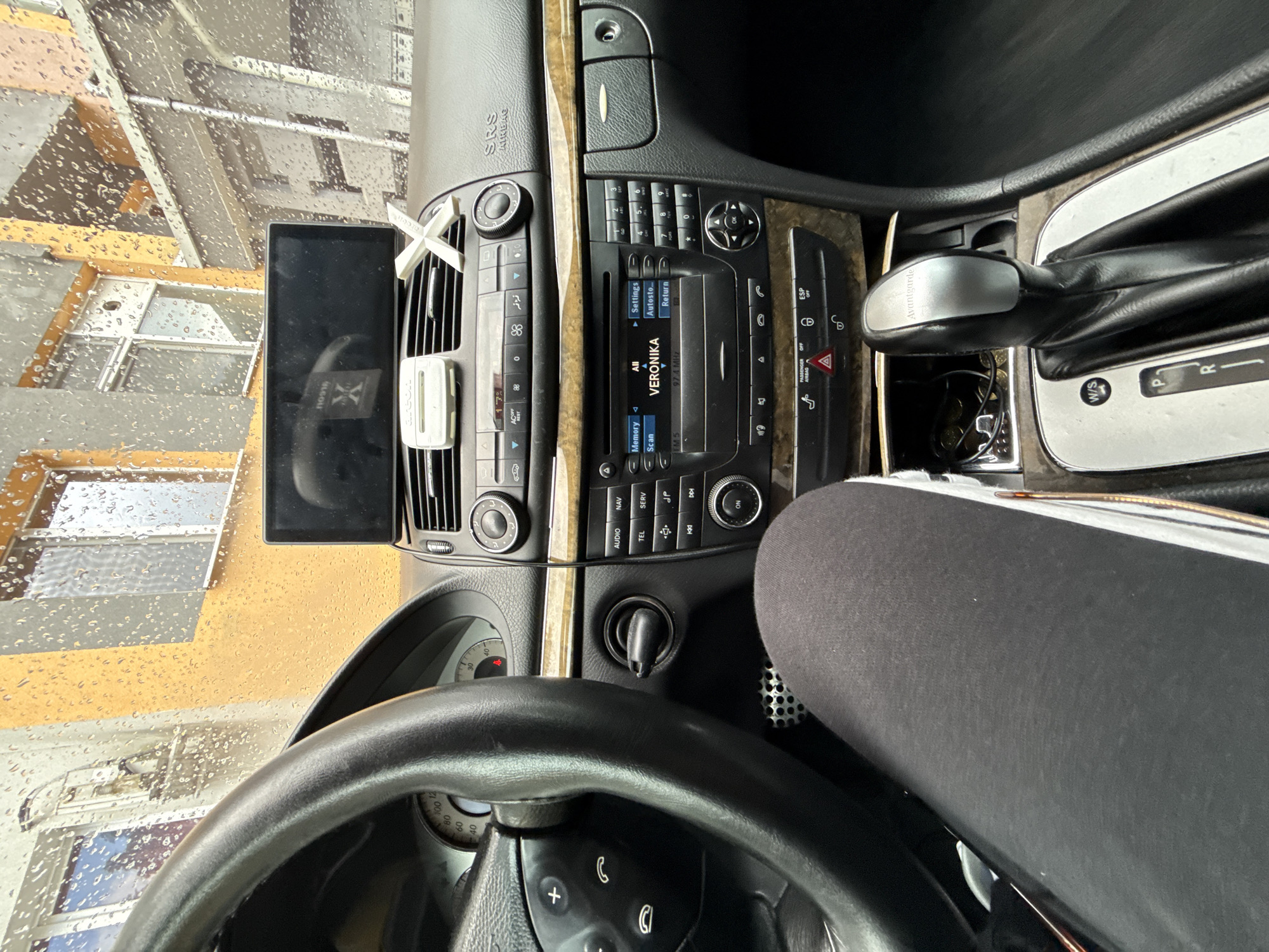 The image captures the interior of a left-hand drive Mercedes-Benz E-Class (W211 generation, indicated by dashboard and gear shifter design, including "Avantgarde" trim on the shifter knob), viewed from the driver's perspective, looking towards the dashboard and out through a rain-splattered windshield. The car is equipped with an automatic transmission and features wood trim accents on the dashboard. An aftermarket widescreen display is mounted above the central air vents, reflecting a stylized white "X" symbol. To the left of this screen, an "AREON" branded air freshener or holder is clipped to an air vent. A separate white "X" shaped ornament is placed on the dashboard to the right of the screen.
The car's integrated infotainment system display shows "VERONIKA" selected, with "97.4 MHz" indicating the current radio frequency. Buttons for "AUDIO," "NAV," "TEL," and "SERV" are visible. Below the climate controls, a hazard warning light is illuminated, and buttons for "PASSENGER AIRBAG OFF" and "ESP OFF" are present. A person's left leg, clad in black pants and white-soled shoes, is visible on the lower left of the frame. A compartment near the gear shifter contains coins and cables.
Outside the vehicle, a multi-story building with an orange/ochre upper facade and a grey lower section is visible through the rain-covered windshield. The weather is heavily raining, causing numerous water droplets to obscure and distort the external view.
The location is the city of Stara Zagora, Bulgaria. This is supported by the presence of a Bulgarian brand "AREON" and the "VERONIKA" radio station, which operates in Bulgaria, although the displayed frequency of 97.4 MHz is typically associated with other Bulgarian cities like Plovdiv for Radio Veronika, rather than Stara Zagora's 96.9 MHz. The architecture of the building outside is consistent with urban environments in Bulgaria.