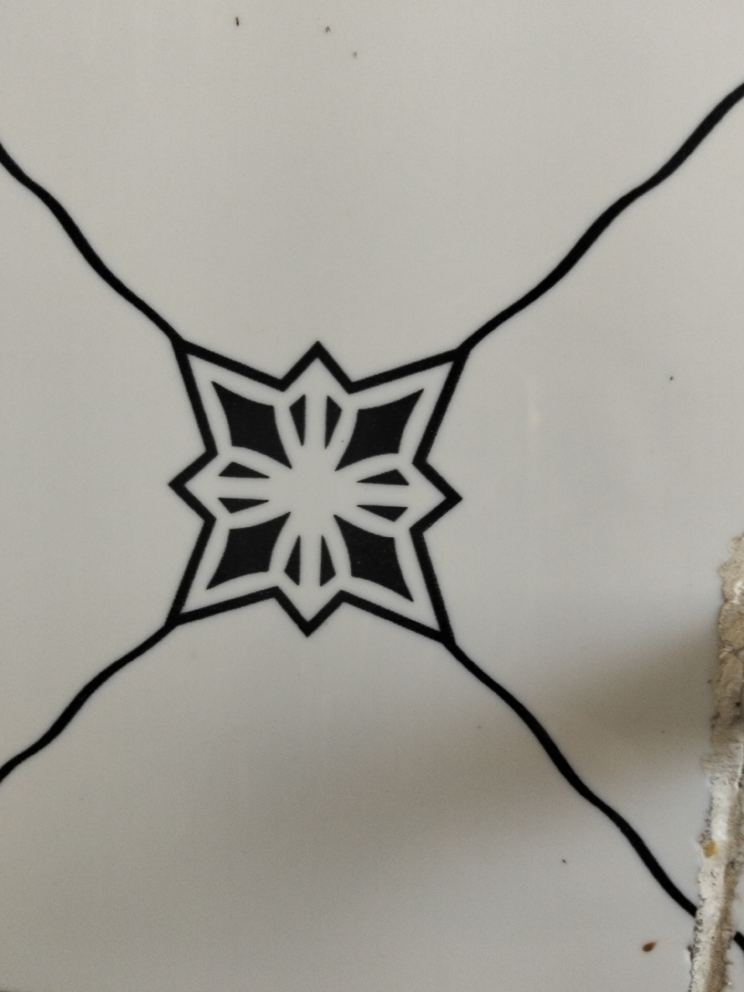 A close-up, flat lay shot of a white tile with a black geometric star design. The design is centered in the frame and consists of a stylized eight-pointed star with radiating lines extending outwards. The tile appears to be part of a larger surface, possibly a wall or floor, given the presence of a rough, damaged edge on the right side of the image, which exhibits a sandy, off-white color and some small brown specks, suggesting it may be plaster or grout. The background is a plain white surface. The image is taken in Kogon City, Uzbekistan.