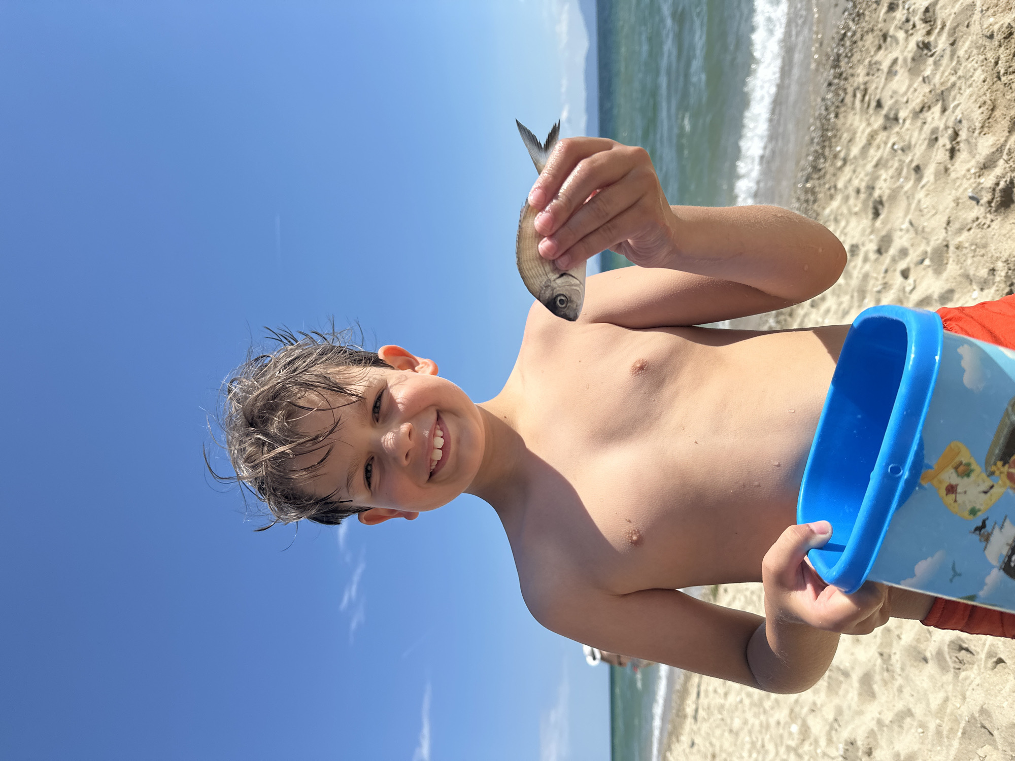 A young boy is smiling at the camera on a sandy beach, holding a small fish in his right hand and a blue bucket in his left. He is shirtless, wearing red shorts, and has wet hair. The ocean is visible behind him, with gentle waves and a clear blue sky above. The location appears to be Ofrynio Beach, Greece.