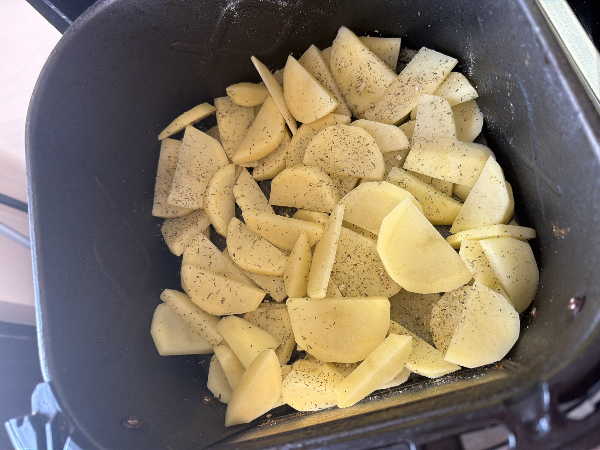 Raw potato wedges, primarily light yellow in color, are shown seasoned and placed inside a dark-colored cooking vessel, likely an air fryer basket or a roasting pan. The potatoes are cut into various shapes, including crescent-like pieces, rectangular chunks, and thinner slices, indicating preparation for cooking. They are visibly coated with dried herbs, which appear as small green and dark brown flakes, and scattered white particles, possibly salt or starch. The seasoning is distributed across most of the potato surfaces. The dark interior of the cooking container features a slightly textured, non-reflective surface, suggesting a non-stick coating. The overall scene depicts food preparation, with the seasoned potatoes ready to be cooked. No people, visible text, time of day, weather, or specific location cues beyond the object types are discernible in the image. The setting appears to be an indoor kitchen or food preparation area.
