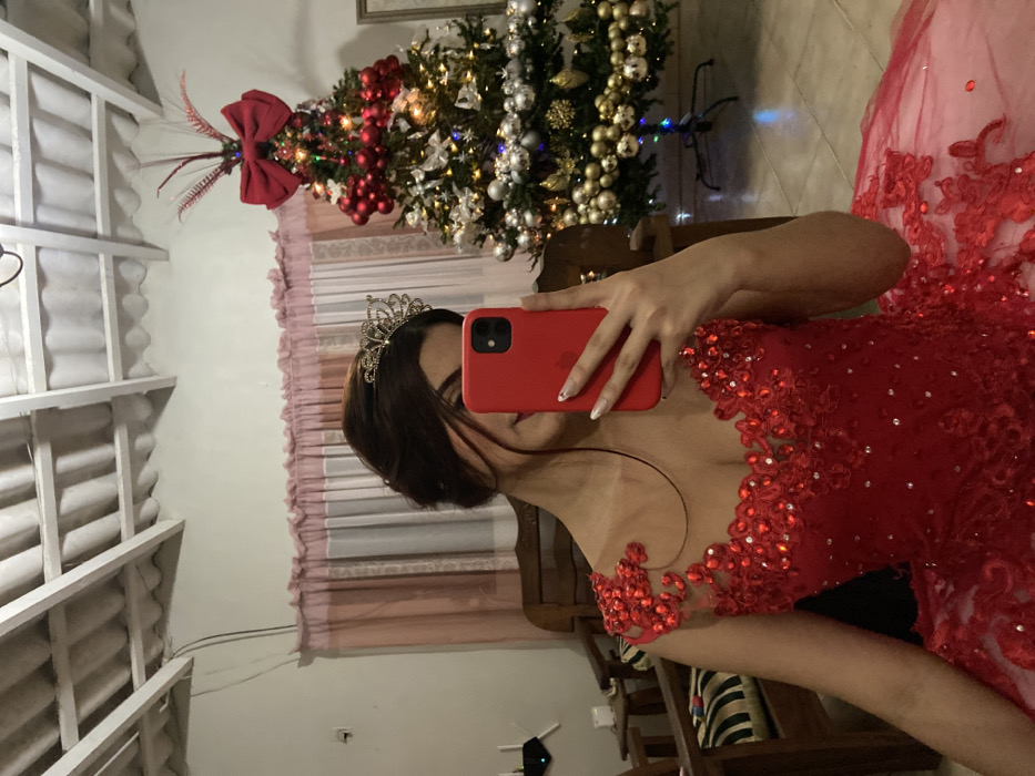 A woman in a red, lace-trimmed gown and a tiara holds a red smartphone, presumably taking a selfie. The background features a decorated Christmas tree adorned with red and silver ornaments, a pink curtain, and a glimpse of a room with wooden furniture. The ceiling, visible in the foreground, is a grid-like structure, possibly a covered patio or porch. The scene is slightly rotated, giving a tilted perspective.