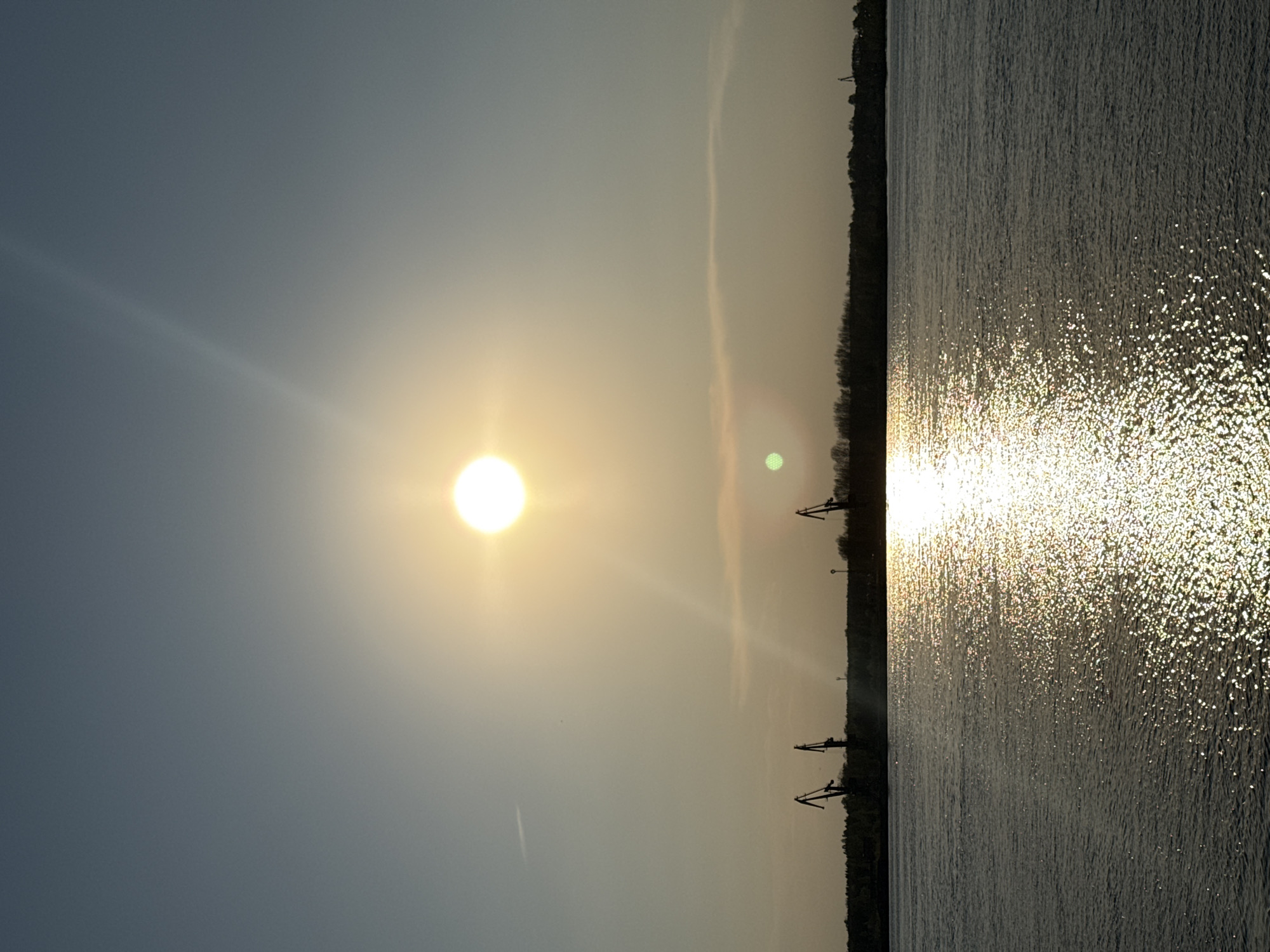 The image shows a body of water reflecting the sunlight at sunset or sunrise, likely in the city of Chaikovsky, Russia. The sun is visible in the sky, with its rays spreading out. On the right side of the image is the water, while the left side shows the sky. Along the horizon, there is a line of trees and some industrial equipment such as cranes visible. The sky is clear, with some thin clouds.