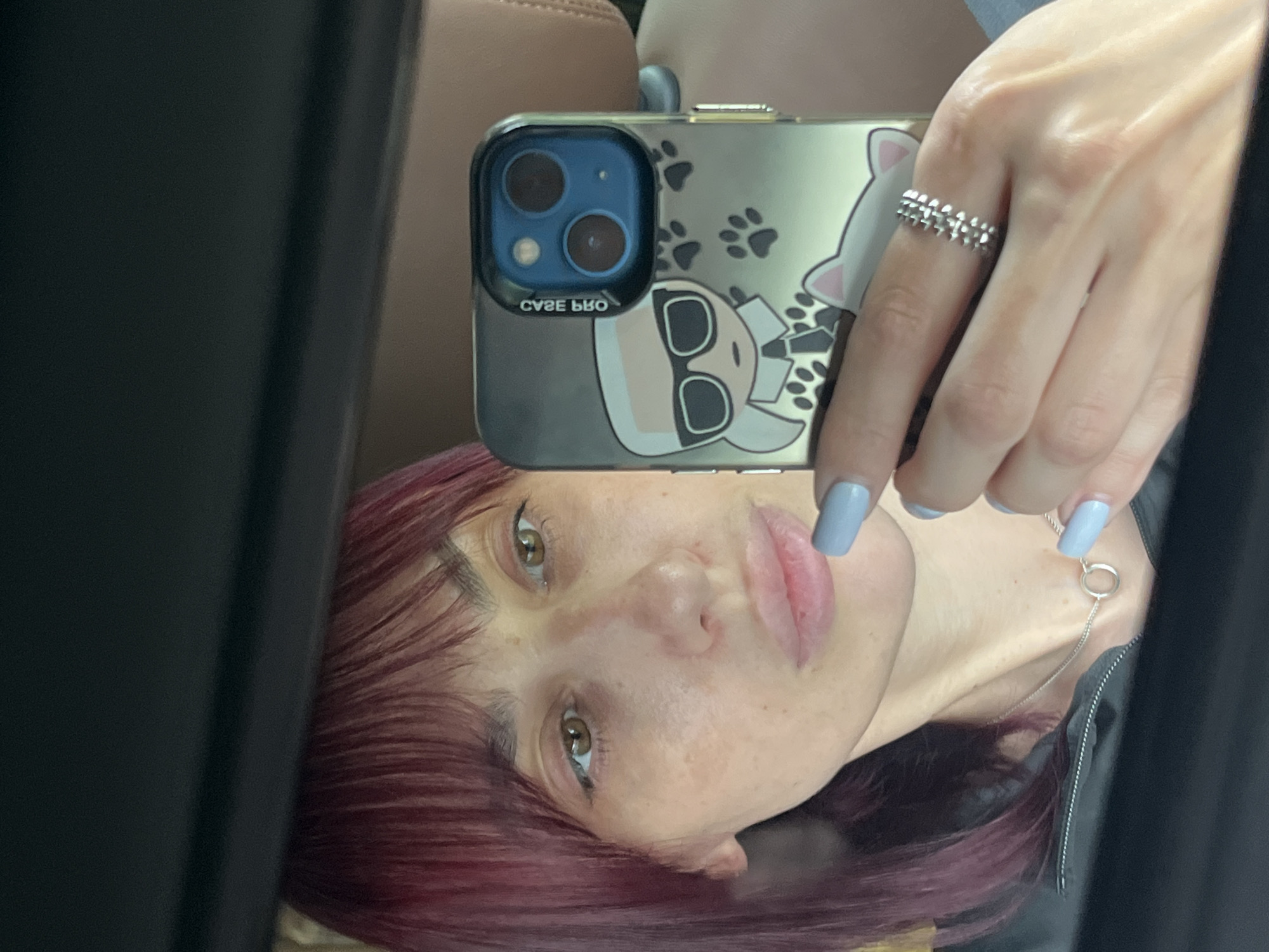The image is a selfie taken in a car, reflected in the car's interior mirror. The primary subject is a person, whose face is visible in the mirror. They have reddish-purple hair, light skin with freckles, and light brown eyes with visible eyeliner. Their lips are slightly parted, and they appear to be looking at the camera.
The person is holding a smartphone, which is also reflected in the mirror, showing the back of the phone. The phone has a clear case with a design featuring a cartoonish character that resembles Karl Lagerfeld, complete with sunglasses, and paw prints. The phone's camera lenses are prominent. The person is wearing a silver ring with a multi-layered, textured design on their right ring finger. Their fingernails are painted a light blue color. A thin silver necklace with a small circular pendant is visible around their neck.
The setting appears to be the interior of a car, with a brown leather seat visible in the background above the person's head. The lighting suggests it is daytime, though no specific time or weather cues are present. The overall mood is casual. No visible text other than "CASEKOO" written on the phone case. The location is specified as Nizhny Novgorod, Russia.