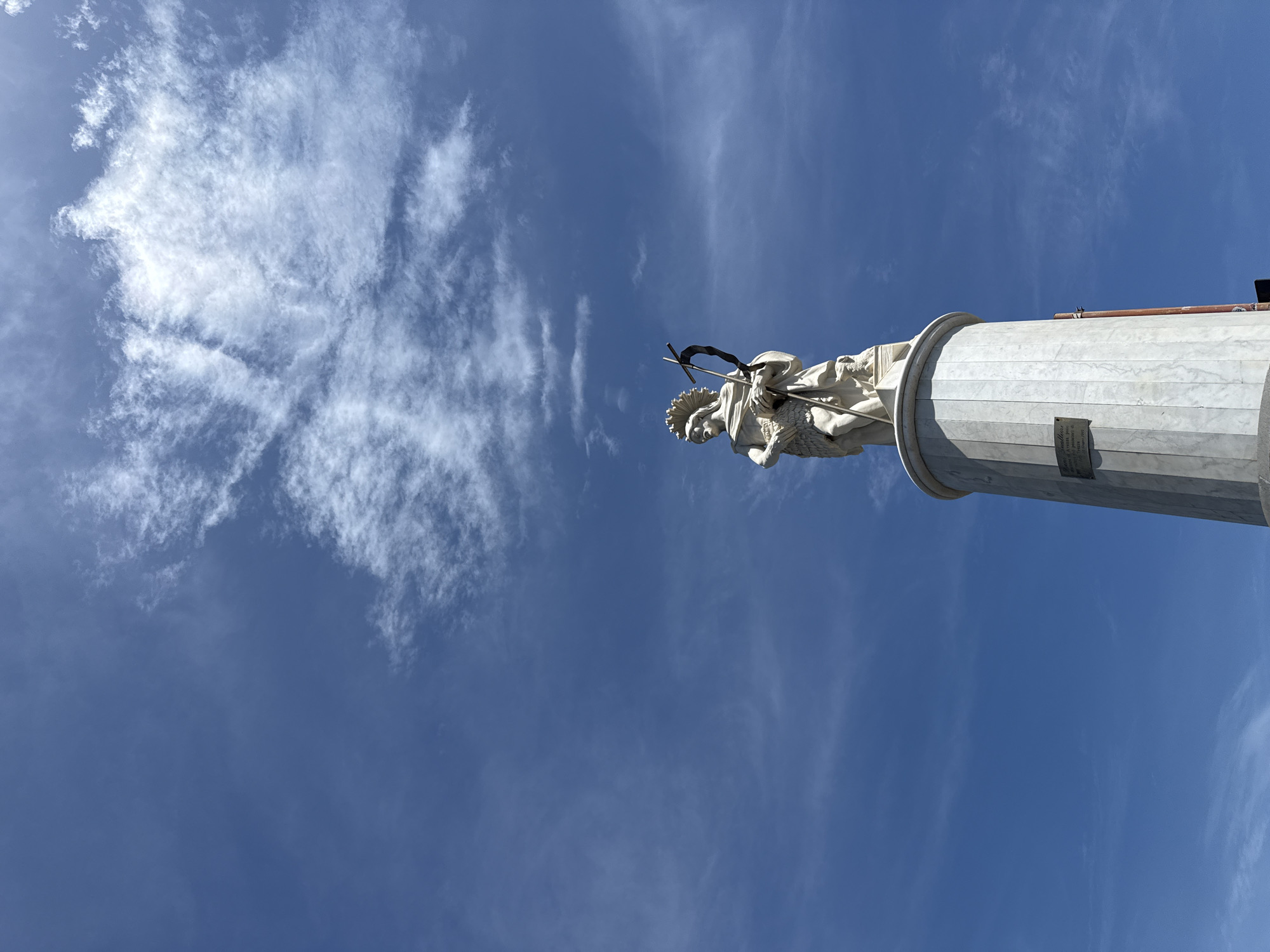 A low-angle shot presents a white statue atop a white column against a partly cloudy blue sky. The statue depicts a figure holding a cross. The sky features prominent cloud formations in the upper-left. The bottom of the column is the bottom-right of the image. A plaque appears near the top of the column, but the text is unreadable. The location is the city of Unknown, Unknown.