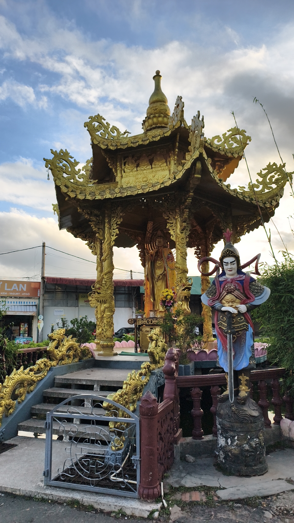 In the city of Xuân Thành, Vietnam, a golden, ornate pavilion stands as the central focus of the image. The pavilion, supported by intricately carved dragon-shaped pillars, houses a large golden statue of a seated figure, possibly a deity. In front of the pavilion, to the right, a colorful statue of a warrior in traditional armor stands guard, holding a sword. The steps leading up to the pavilion are adorned with golden dragon railings. The scene is set against a backdrop of a cloudy sky, with hints of a nearby building and vegetation visible. A metal gate in the foreground partially obscures the lower steps, suggesting a public or accessible area. The overall impression is one of cultural significance and artistic craftsmanship.