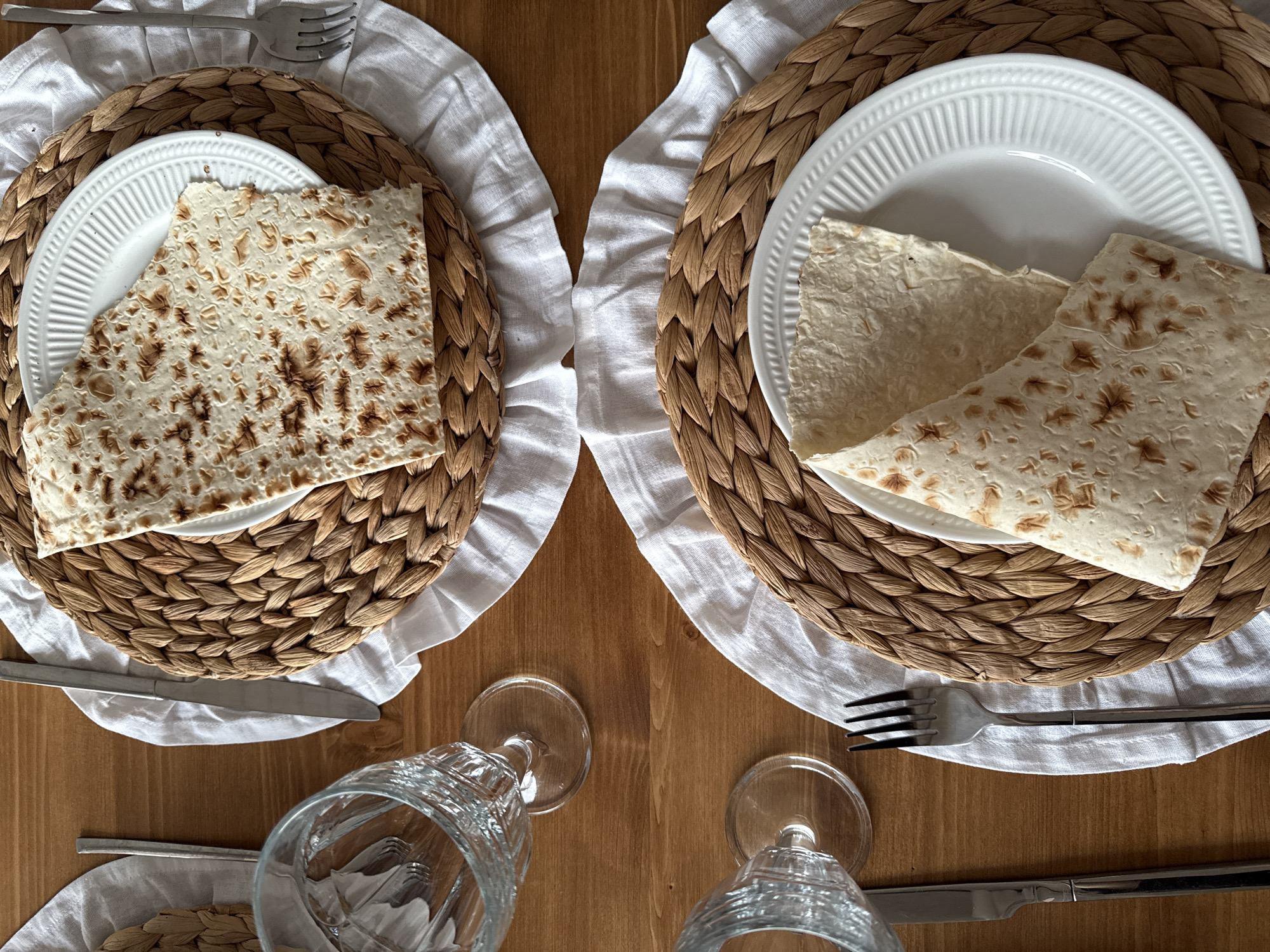 A top-down, medium shot captures a wooden table set for two with plates of matzah. Each place setting includes a woven placemat, a white linen napkin, a white plate, silverware, and a glass. Two clear glasses are partially visible on the left side of the image. The matzah on the plates is flatbread, some pieces folded and others laid flat. The lighting is natural, creating soft shadows. The scene appears to be in a home setting in Starokorsunskaya, Russia.