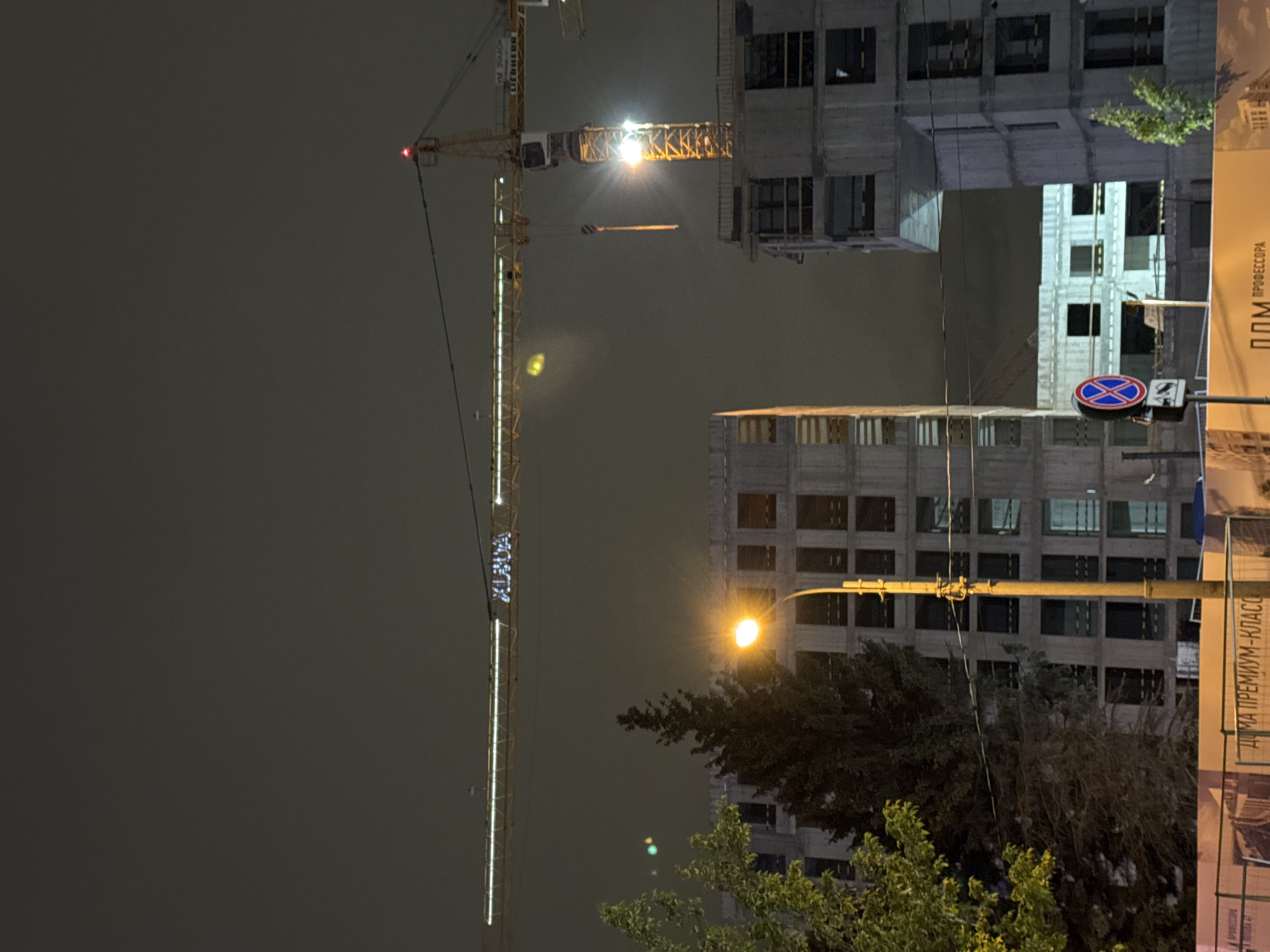 The image depicts a nighttime scene in Saint Petersburg, Russia, featuring a construction site. A tall crane, partially illuminated, is prominent in the view, alongside a building under construction. Streetlights are also visible, casting a warm glow. A street sign with a "no stopping" symbol is visible. Trees partially obscure some of the buildings. The general atmosphere is urban and related to construction activities. The text on a sign reads "ДОМА ПРЕМИУМ-КЛАССА".