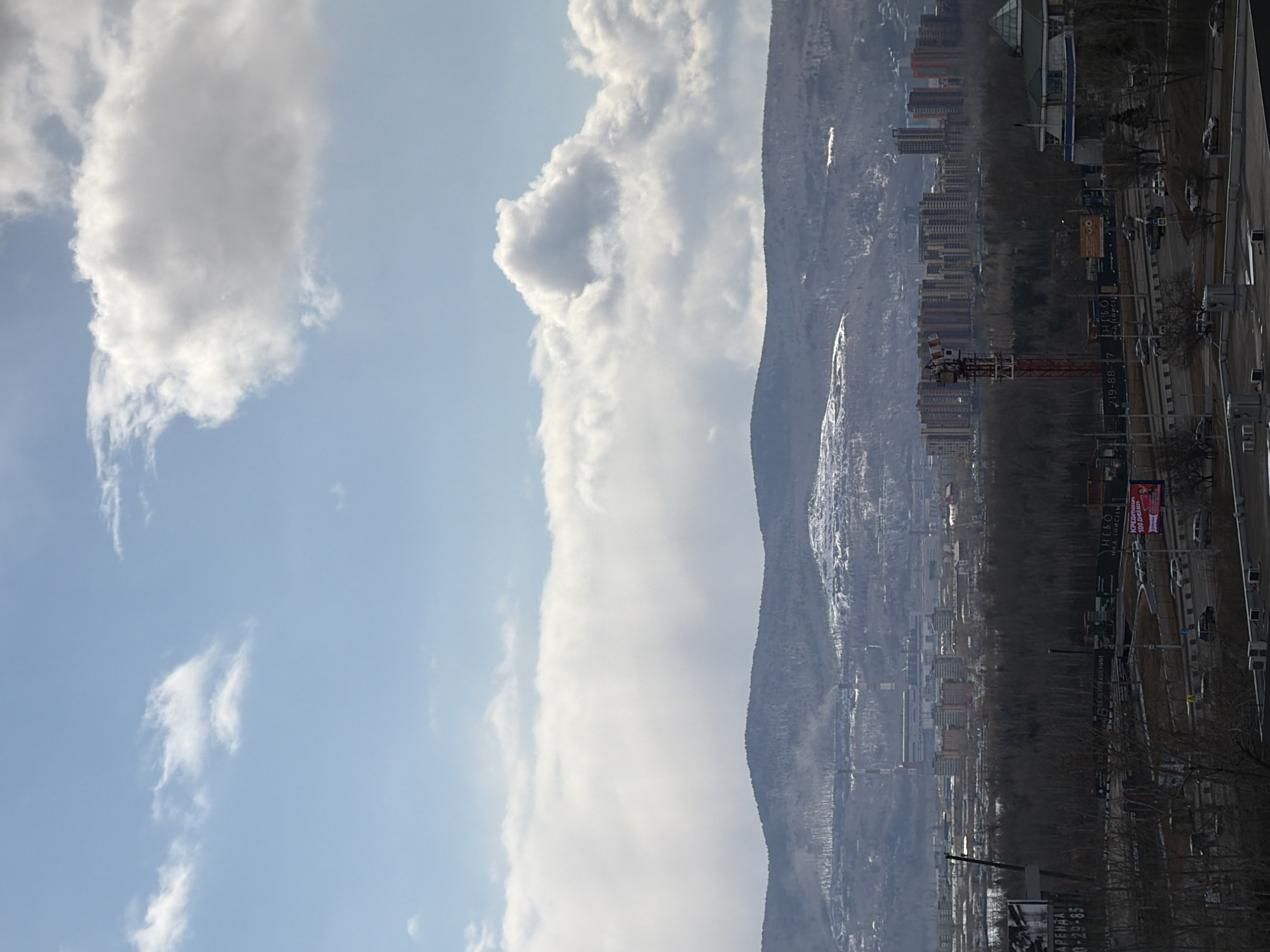 The image presents a view of Krasnoyarsk, Russia. The right side of the frame shows a cityscape with apartment buildings, commercial buildings, roads with moving vehicles, and infrastructure elements like street lights and signs. A construction crane is visible amidst the buildings, indicating ongoing development. The base of a mountain with snow is behind the city. The left side of the frame is dominated by a blue sky with white clouds. The image appears to have been taken from an elevated position.