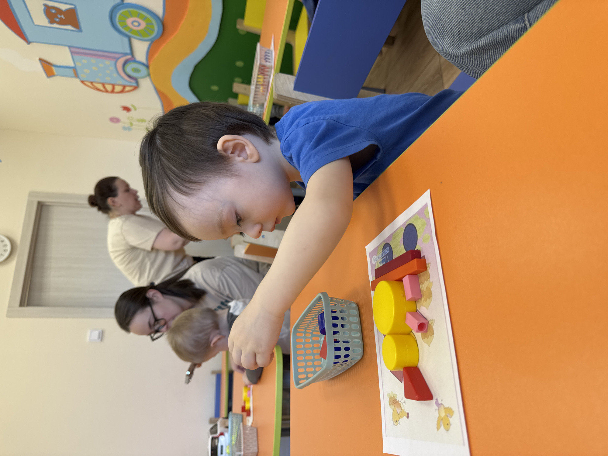 A child in a blue shirt is at a table with a shape-sorting activity in Izhevsk, Russia. There is a white sheet with shapes outlined on it and a basket of shapes. Other children and adults are present in the background. The walls have colorful murals of a train.