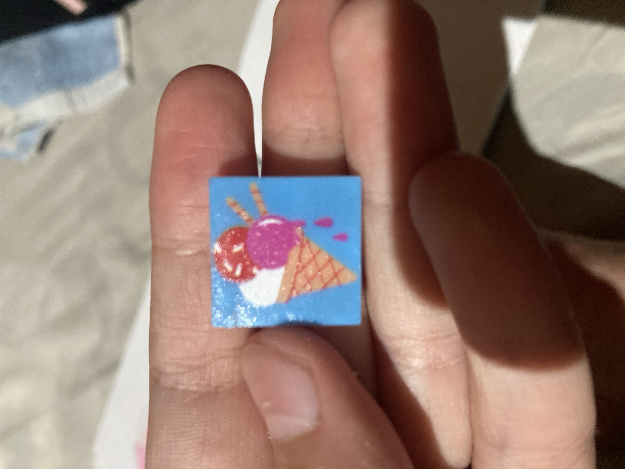 A person is holding a small, square piece of paper with a colorful image of an ice cream cone against a bright blue background. The ice cream has three scoops: one red with sprinkles, one pink, and one white. The cone is a light brown waffle cone with orange cross-hatching. There are two pink drips of melted ice cream falling from the cone. The background is a blurred light gray, possibly a bedspread or fabric. The person's fingers are visible, gently supporting the paper square. The image does not depict any specific location in the city of Besedy, Russia.