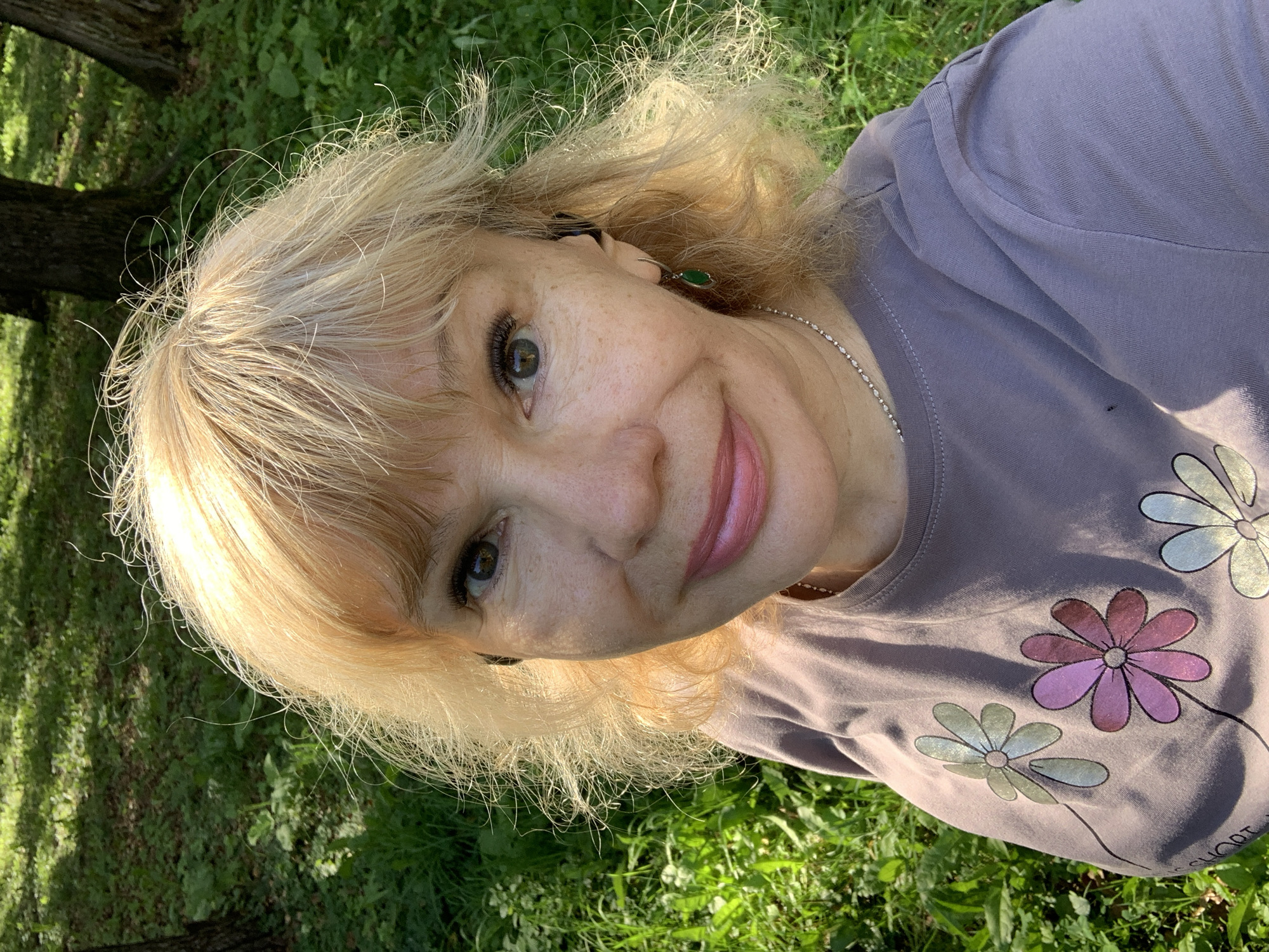 An outdoor, waist-up, high-angle selfie shows a fair-skinned woman with blonde, windswept hair and light freckles on her face and chest, posing in a green, sun-dappled park with trees and grass. She is wearing a lavender t-shirt with a floral design of pink, purple, and light green flowers, and a delicate silver necklace. She has on small, dark earrings, one visible on her right earlobe with a dangling green stone. She is also wearing what appear to be wireless earbuds. The woman is looking directly at the camera with a slight smile and her eyes are open. The lighting suggests it is daytime, likely in the afternoon, with dappled sunlight casting shadows on the grass. The setting is a natural, outdoor environment consistent with a park or wooded area. The location is the city of Prokopyevsk, Russia.