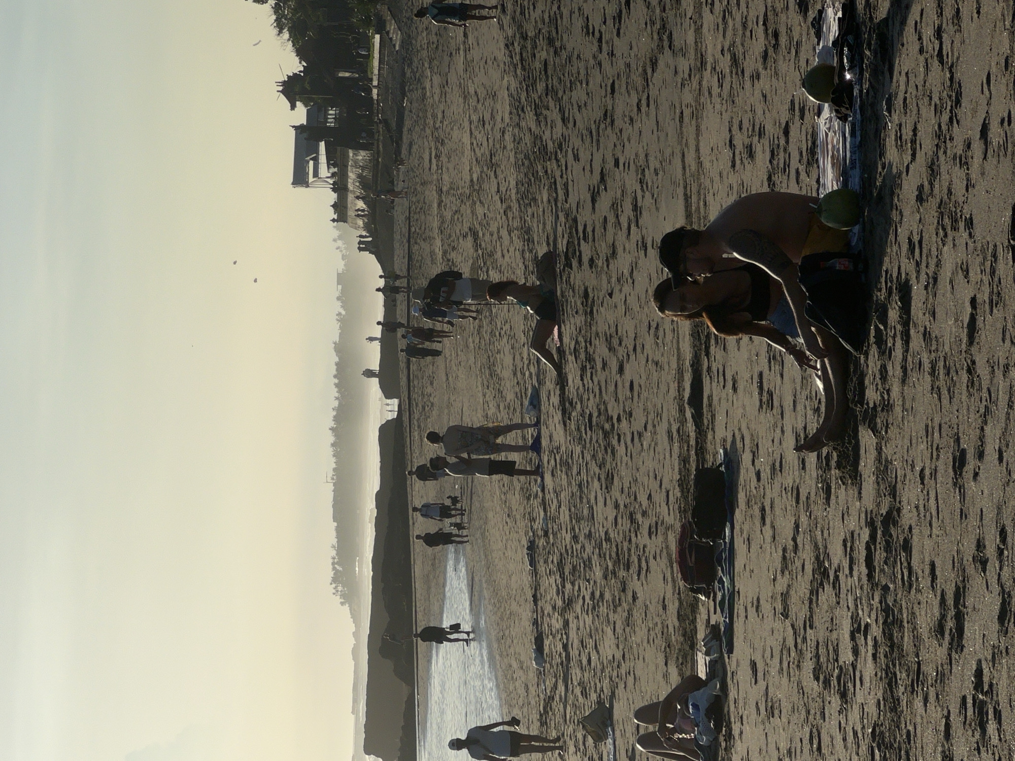 The image depicts a hazy beach scene in North Kuta, Indonesia, likely during sunrise or sunset, indicated by the warm, atmospheric light and long shadows stretching across the dark sand. The beach, possibly composed of volcanic sand, features wet areas reflecting the soft light. In the distance, a coastline lined with palm trees and silhouettes of traditional Indonesian buildings or temples is visible through the haze.
Numerous people are scattered across the expansive beach. In the foreground, a couple is embracing intimately, largely silhouetted against the bright horizon. The man wears a cap and yellow shorts, revealing an arm tattoo, while the woman is in a dark bikini top and light-colored shorts. Nearby, two green coconuts and a patterned towel or mat rest on the sand.
In the midground, several individuals and small groups are seen walking, standing, or bending over, some wading in the shallow water at the edge of the receding tide. One person appears to be carrying a paddleboard or surfboard. Other personal items such as bags and shoes are visible on the sand. The overall atmosphere conveys a sense of leisure and enjoyment of the tranquil time of day. No visible text is present in the media file.