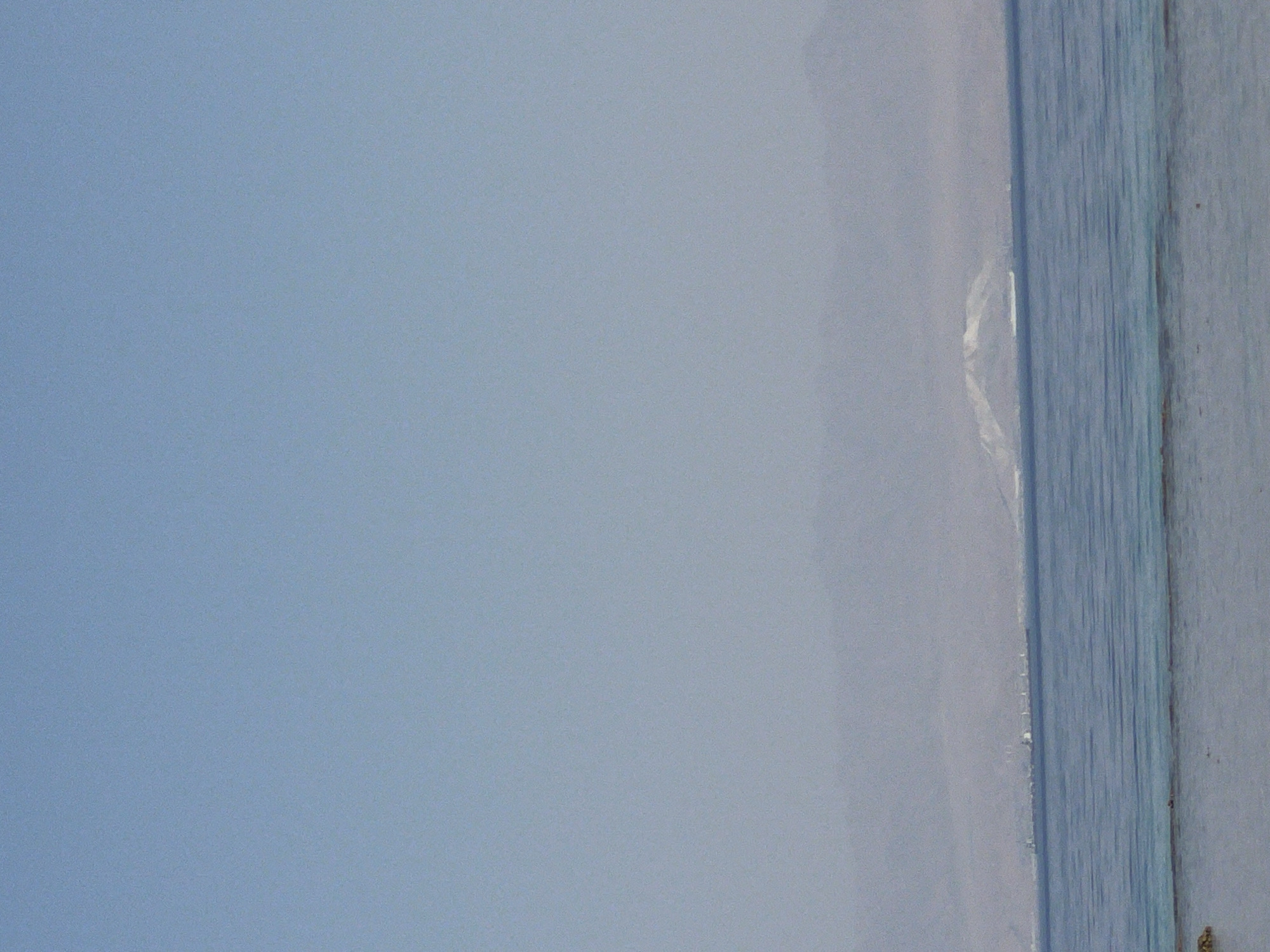 The image depicts a panoramic view of the sea and distant land under a clear blue sky. The foreground shows shallow, turquoise water with a sandy bottom visible. Beyond this, a wider expanse of deeper blue water stretches to the horizon. In the middle ground, a line of hazy mountains forms the distant coastline. A prominent, snow-capped mountain peak is visible in the center of the mountain range, suggesting it could be Mount Sinai or a related peak, indicating the location is likely Nueiba, Egypt, which is situated near these mountains. Several ships, appearing as white specks on the water, are present in the distance, possibly in a shipping lane or port. The overall atmosphere is serene and vast, characteristic of a coastal landscape.