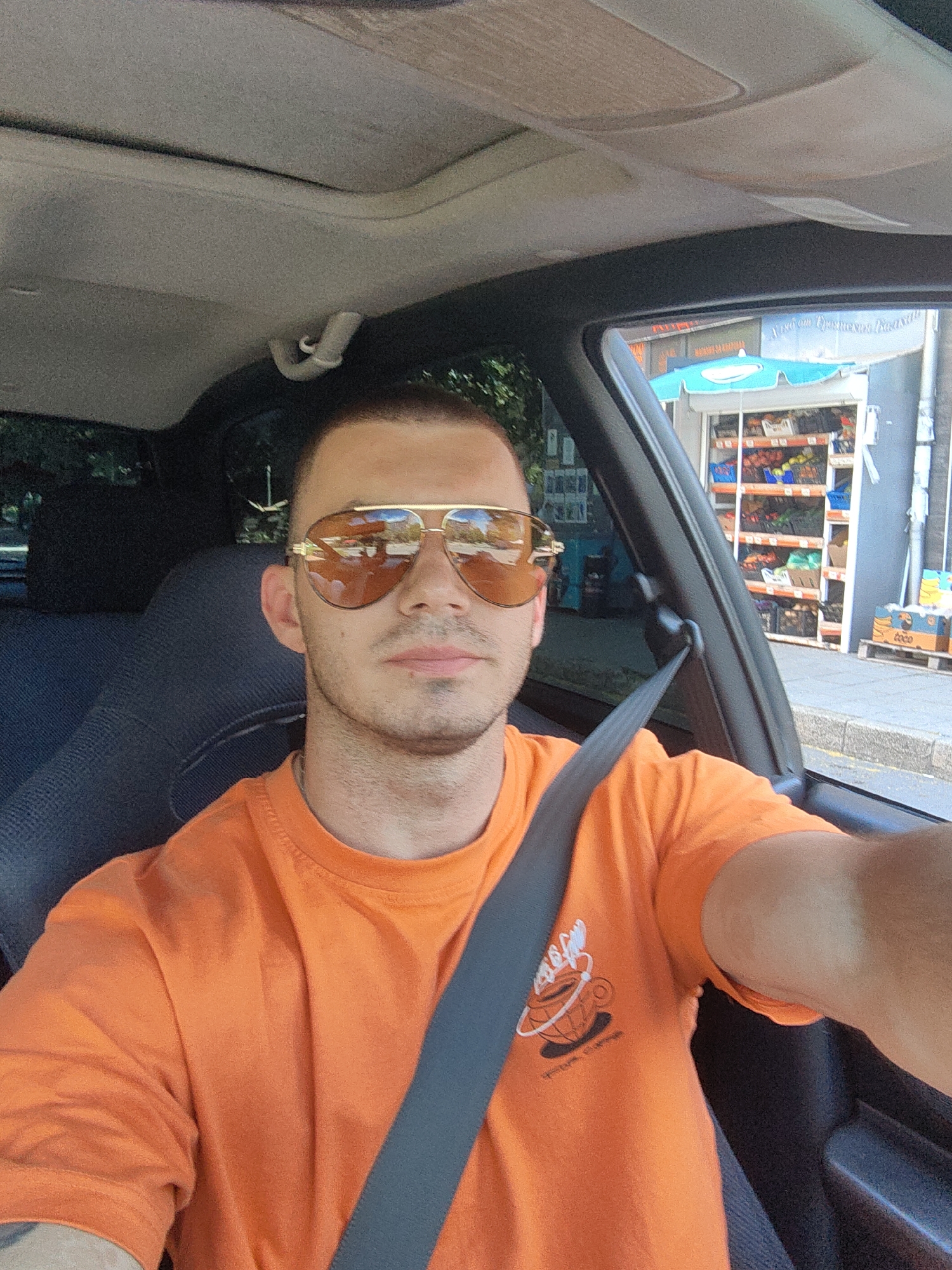 A man is taking a selfie while sitting in the driver's seat of a car. He is wearing an orange t-shirt with a graphic of a coffee cup and steam. He has on gold aviator sunglasses with amber lenses that reflect the surroundings. The interior of the car is visible, including the grey seat and a portion of the car's headliner and sunroof. The man is looking at the camera and appears to be in a neutral or slightly smiling mood. He has short hair and stubble. A black seatbelt is fastened across his chest.
Through the car window on the right side, a street scene is visible. There is a shop with shelves displaying various products, likely produce, and a light blue umbrella over an entrance. There are also some crates and pallets outside the shop. The background suggests an urban environment with buildings and trees. The time of day appears to be daytime, with bright sunlight casting reflections. The weather seems to be fair.
The location is Varna, Bulgaria, and the visible text on the shop sign is in a language with Cyrillic characters, consistent with Bulgaria. One visible text on a crate reads "toco." The graphic on the t-shirt also has some text, but it is not clearly legible.