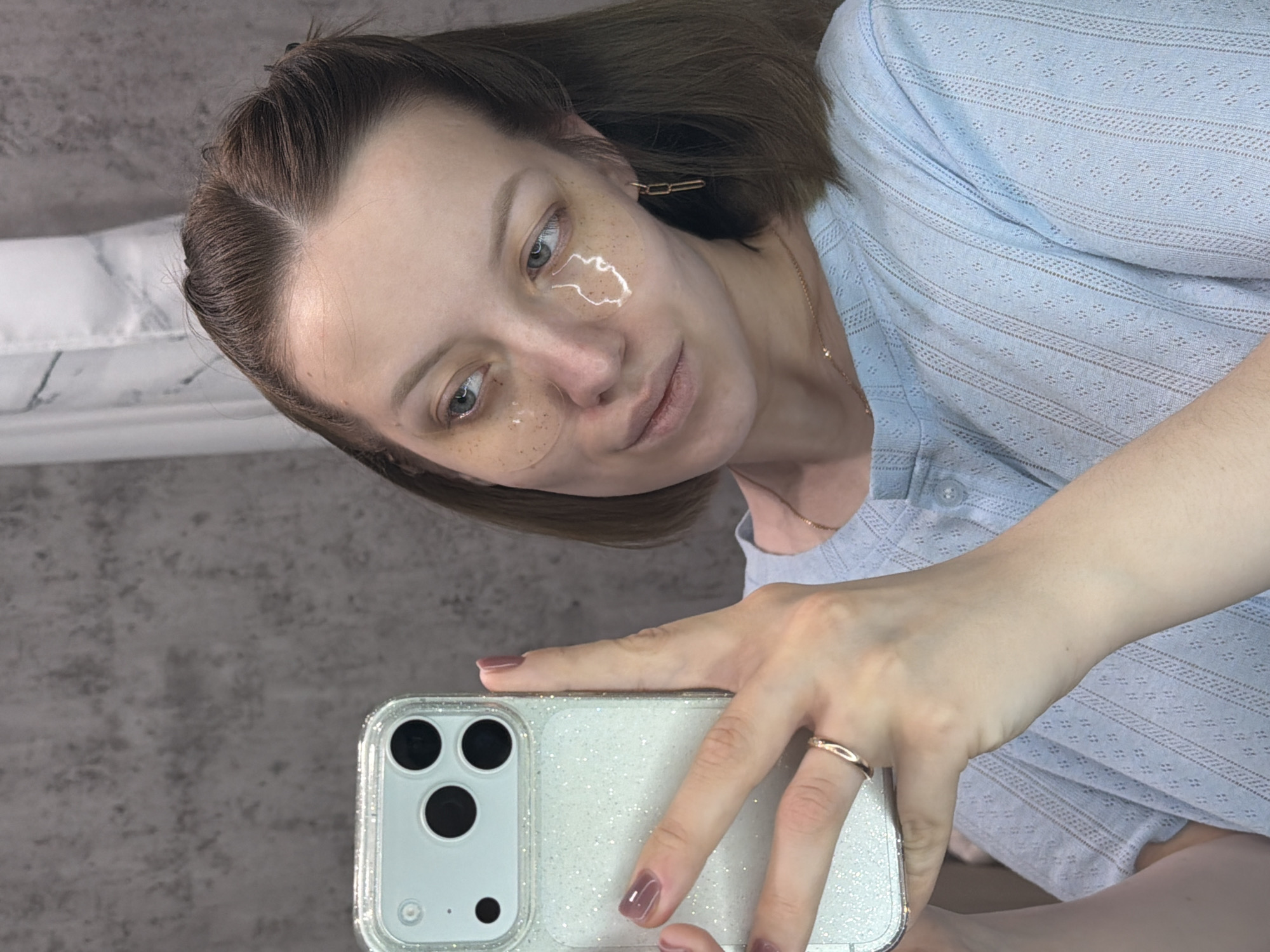 A woman takes a selfie indoors, positioned at a slight angle. She has dark brown hair pulled back, revealing a clean forehead, and her eyes are a light greenish-blue. On her cheeks and under-eye area, she wears clear hydrogel patches containing small flecks, indicative of a skincare routine. Her expression is neutral and calm as she looks towards the camera.
She is dressed in a light blue, short-sleeved top with a textured pattern, possibly eyelet or embroidered, and wears a delicate gold-toned necklace. On her right hand, which holds the smartphone, she has a simple gold-colored ring on her ring finger, and her fingernails are painted a muted purplish-brown hue.
The smartphone she holds appears to be an iPhone with three camera lenses, housed in a glittery clear case. The background consists of a light gray, textured wall and, partially visible to her left, a white pillow with a subtle marble pattern. The lighting is soft and even, suggesting an indoor setting. There is no visible text in the image. The overall scene conveys a moment of personal care in a relaxed, home environment.