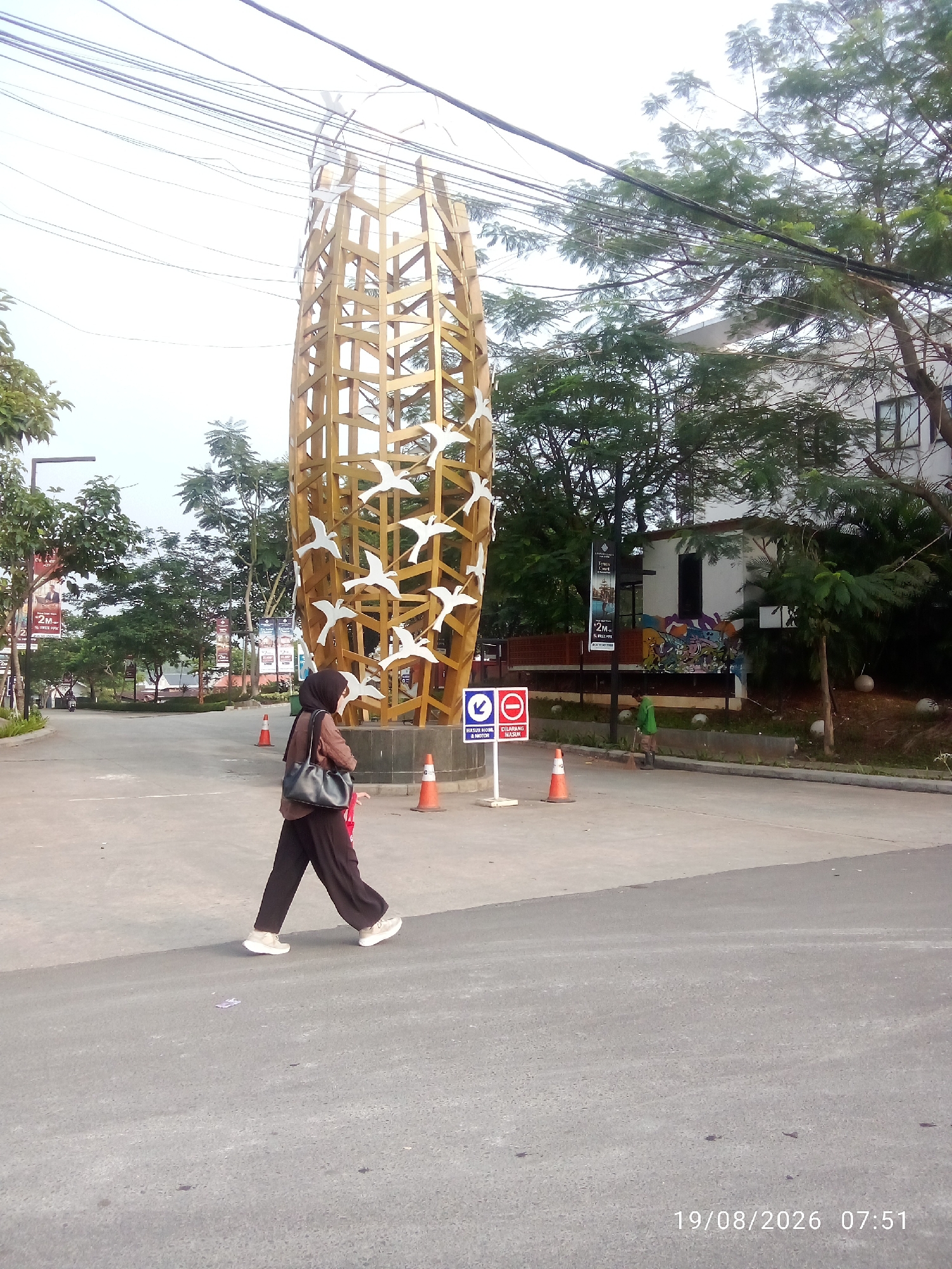 The image captures an outdoor scene in East Jakarta, Indonesia, featuring a large, golden, birdcage-like sculpture adorned with white bird silhouettes. In the foreground, a woman wearing a hijab and dark clothing walks across an asphalt surface, carrying a brown shoulder bag. She is wearing white sneakers. Orange traffic cones are visible on the pavement near the sculpture. In the background, there are trees, a building, and banners. A person in a green hooded jacket is seen sweeping the ground near a small structure with graffiti. A sign with a blue arrow pointing left and a red circle with a diagonal line across it, indicating "No Entry," is positioned on the left side of the image. The timestamp "19/08/2026 07:51" is displayed in the lower right corner.