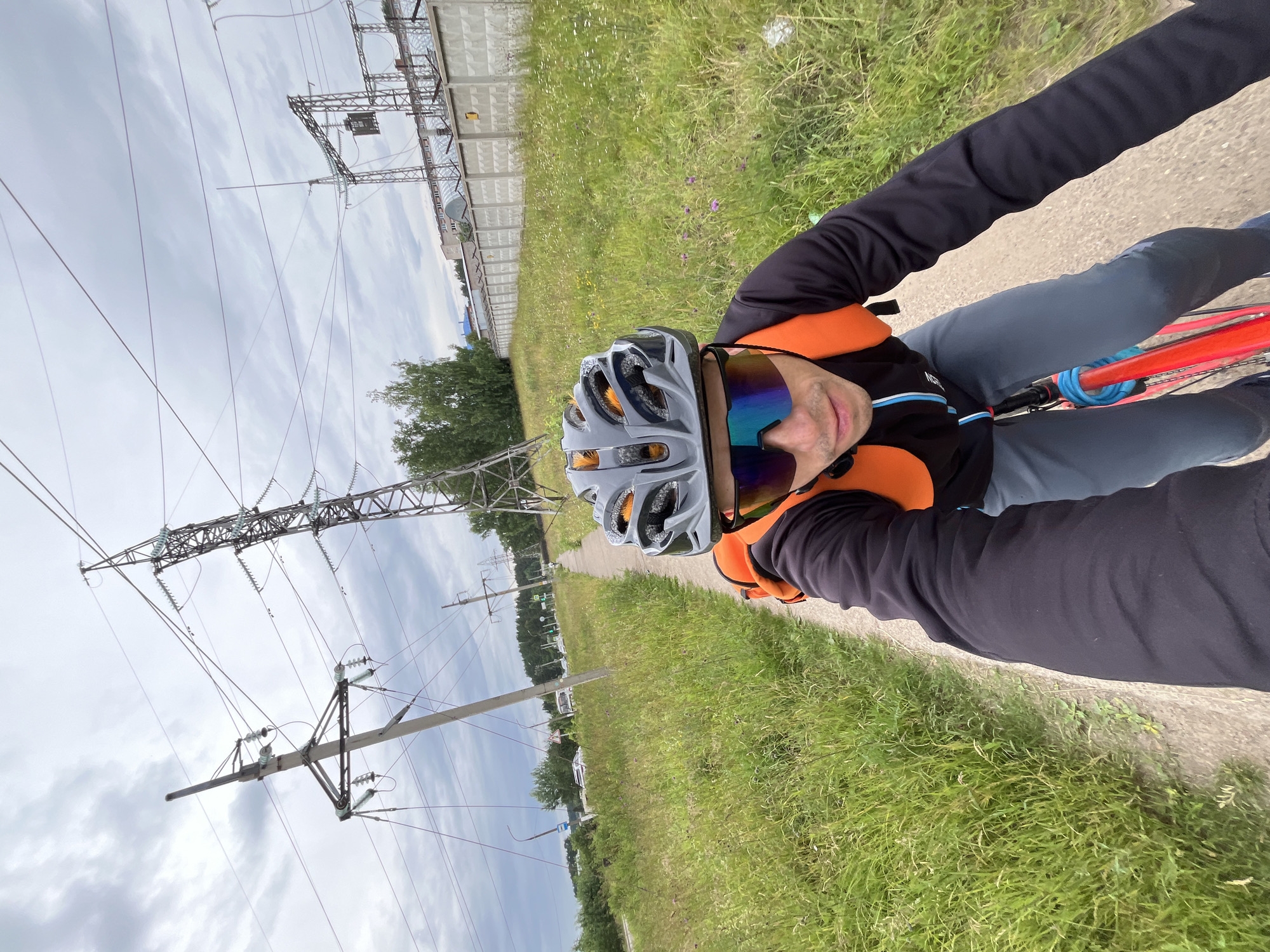 An individual wearing a helmet, sunglasses, a black long-sleeved shirt, and grey pants is cycling on a dirt path. They have an orange backpack on. The path is bordered by tall green grass and wildflowers. In the background, there are large power lines and a concrete fence. Beyond the fence, there are some buildings and trees. The sky is overcast with grey clouds. The location is likely in the city of Chaikovsky, Russia, given the presence of industrial infrastructure like power lines and a substation.