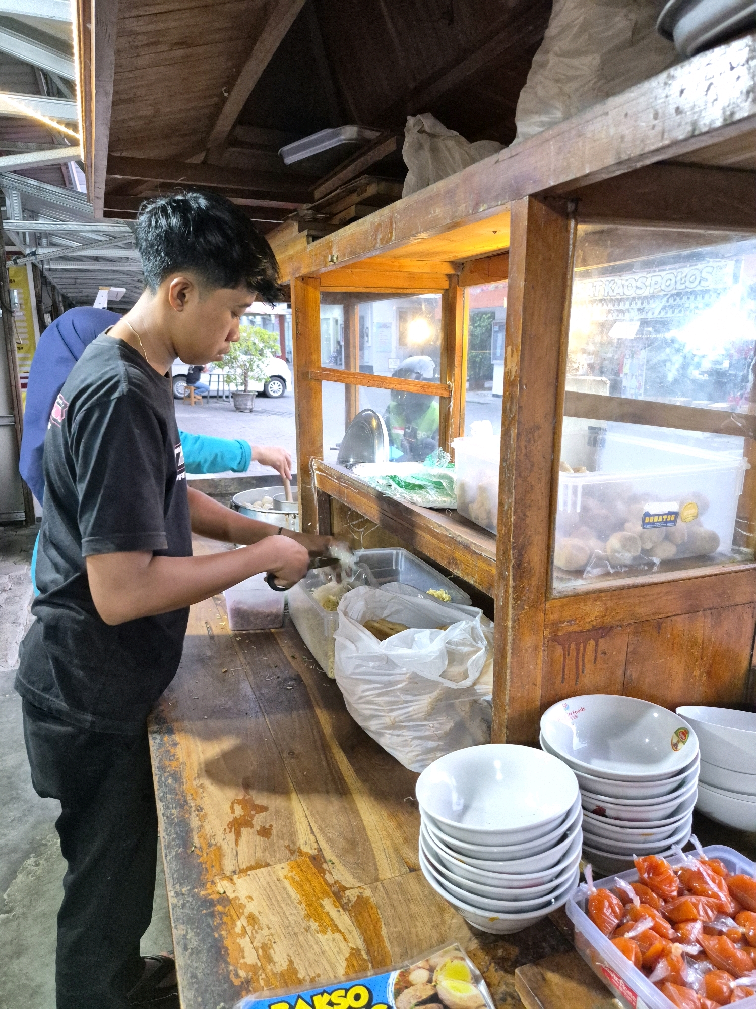 The image depicts a street food vendor preparing food at a wooden cart in Purworejo, Indonesia. A young man, wearing a black t-shirt and black pants, is actively engaged in preparing ingredients, holding a pair of scissors in his right hand and working with some shredded food items. He has dark, short hair and is facing the cart. Behind him, partially visible, is another person wearing a blue shirt and blue hijab, also attending to the food preparation. The cart itself is made of wood and features glass panels displaying food items. Visible items within the cart include plastic containers with what appear to be meatballs or similar food products, and stacks of white bowls. In the foreground, on the wooden surface of the cart, there are more stacks of bowls and a plastic container filled with small, orange-wrapped food items, likely condiments or snacks. The overall environment suggests an outdoor food stall with a rustic, functional setup. The background shows a slightly blurred view of a street scene, possibly with a parked vehicle and trees, indicating an urban or semi-urban setting. The lighting suggests it is either early morning or late evening.