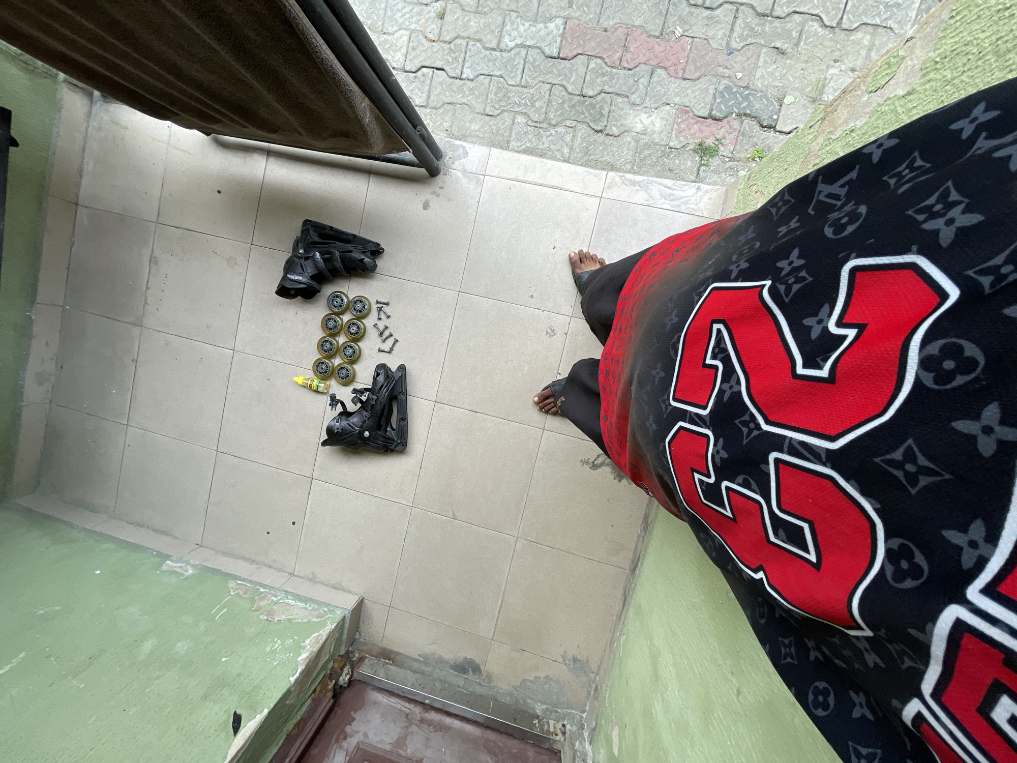 The image depicts an overhead view of a person's legs and feet standing on a tiled floor, wearing black pants and a red and black patterned shirt with large red numbers. In front of the person are disassembled inline skates, including two black boots, several yellow wheels, and a small bottle of lubricant. The scene appears to be outdoors, possibly on a porch or patio, with a green painted wall and a wooden door visible to the left, and patterned paving stones to the right. The overall setting suggests a casual, residential environment in Ado, Nigeria.