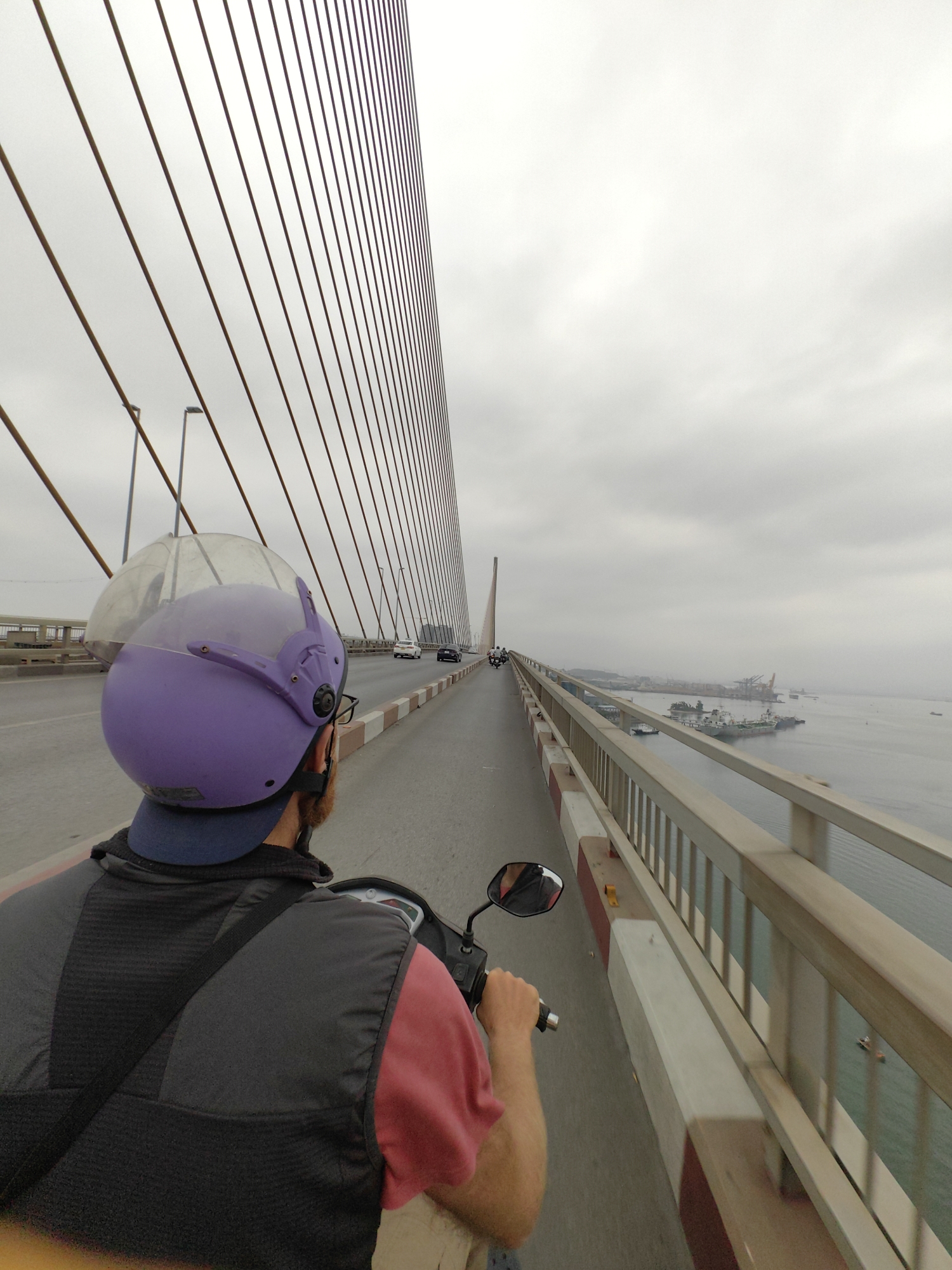 A person is riding a scooter across a suspension bridge, identified as a bridge in Hong Gai Ward, Vietnam, as suggested by the port and industrial facilities in the background. The rider wears a purple helmet and a dark vest over a red shirt. The bridge features a pattern of parallel cables extending from a central tower towards the roadway. The scene is overcast, and the bridge railing is visible on the right side of the frame, with a body of water and ships in the distance.